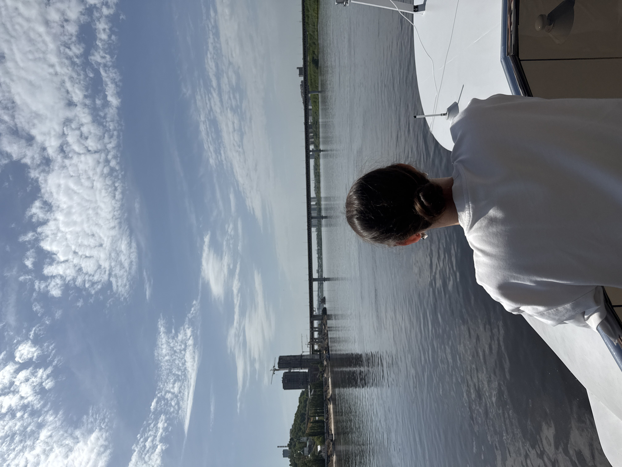 The image shows a view from what appears to be a boat or ship on the Volga River in Nizhny Novgorod, Russia. A woman in a white shirt is standing near the edge of the vessel, overlooking the river. In the background, a bridge spans the Volga, connecting different parts of the city. On the riverbanks, buildings and trees are visible, indicative of the urban landscape of Nizhny Novgorod. The sky is partly cloudy.