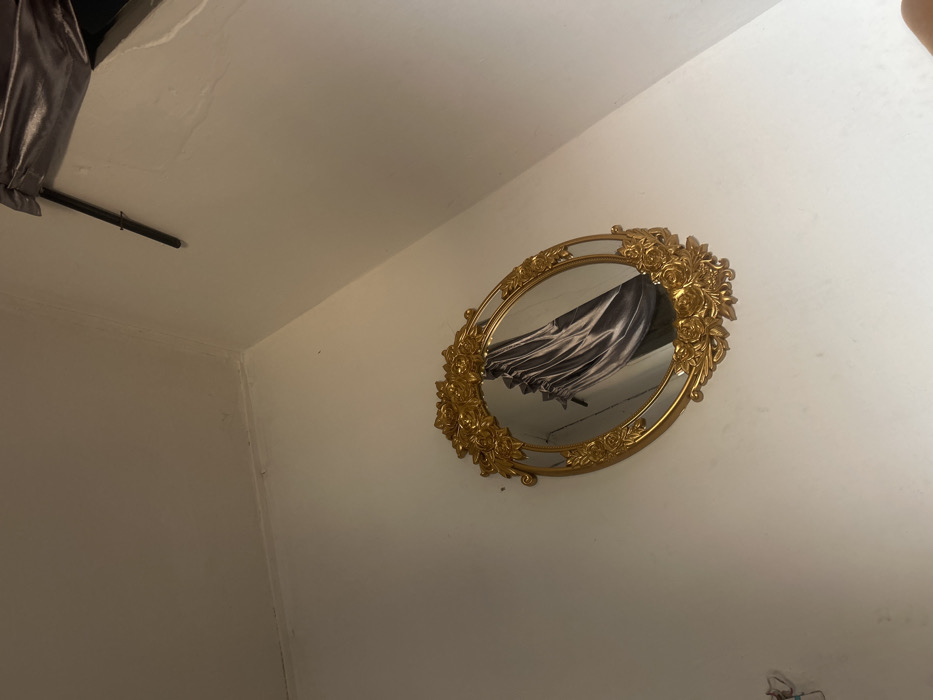 The image depicts an indoor scene in Ilishan, Nigeria, focusing on an oval-shaped mirror with a prominent gold, ornate frame featuring floral motifs, mounted on a plain white wall. The mirror's reflection shows a textured, dark grey or silver fabric, consistent with a drape or curtain, partially obscuring a dark background. The wall surface appears smooth, with a subtle seam or corner visible in the upper left. In the upper right corner of the overall scene, a dark, thin rod is horizontally mounted, partially obscured by a section of dark, metallic-sheen fabric, possibly part of an awning or another decorative element, that is slightly angled downwards from the ceiling. No people are visible. The environment suggests an interior space with minimal furnishings beyond the mirror and the reflected elements.