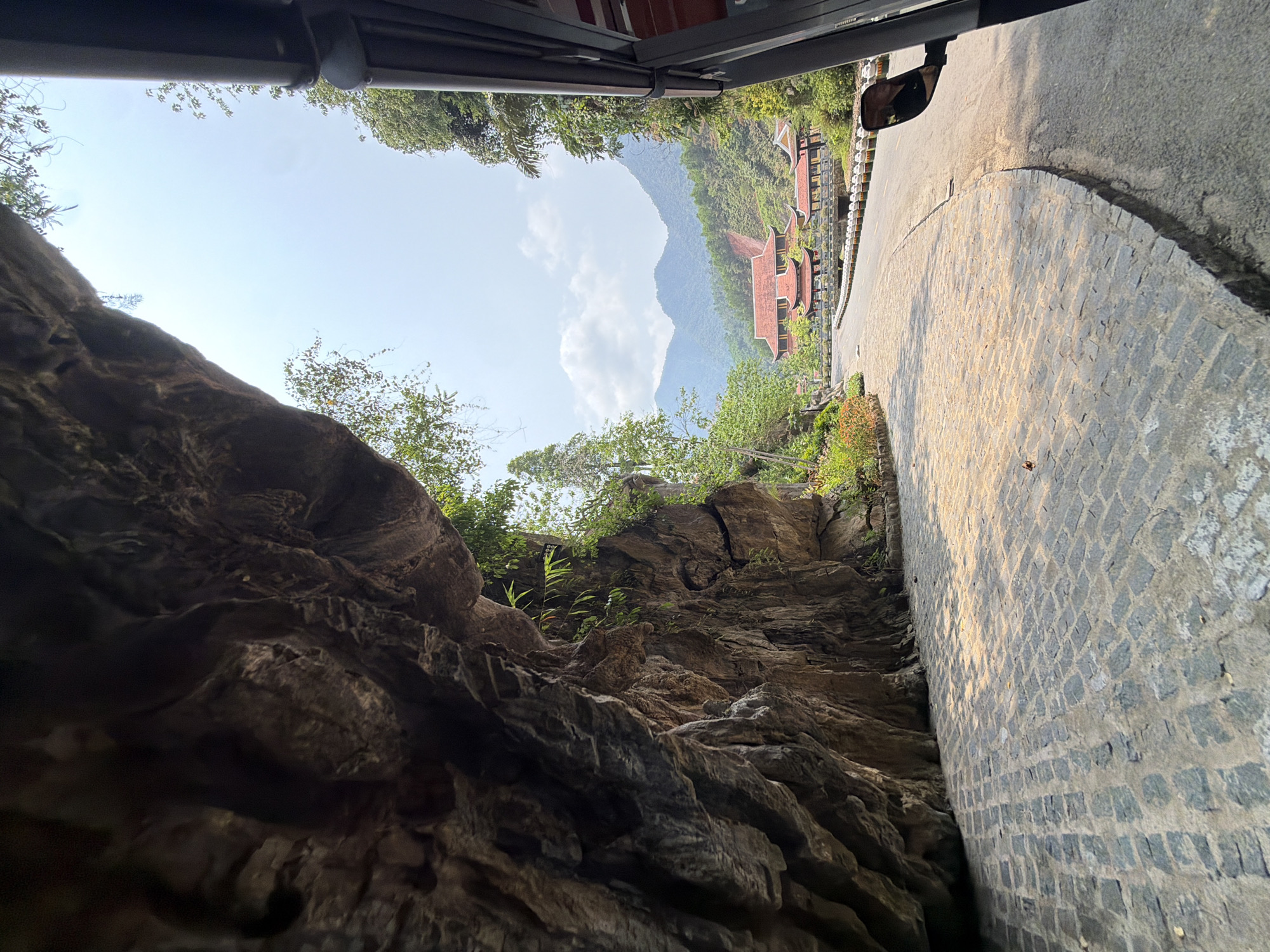 The image, taken from a low angle, shows a narrow space between a large rock formation and a paved road leading to a building with a red roof. The sky is visible at the top of the frame, with some greenery. The image appears to be taken from inside a vehicle, looking out through a small opening.
The location is likely the Marble Mountains in Đà Nẵng, Vietnam, known for its caves, temples, and pagodas, which often have roads winding through rocky landscapes. The red-roofed building is a common architectural style for temples in the area.