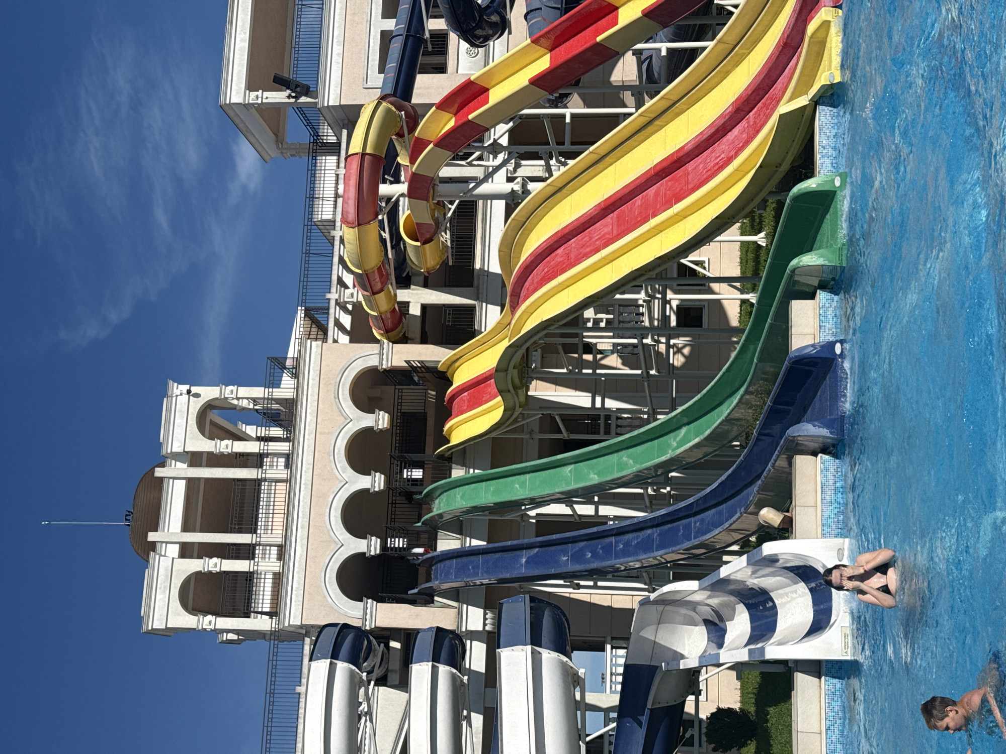 This media file depicts an outdoor water park on a sunny day. The scene is dominated by a large, multi-level water slide structure. The slides are colorful, with sections of red and yellow, green, and blue, twisting and turning as they descend towards a bright blue swimming pool.
In the pool, two young people are visible. One, a person with dark hair wearing a black swimsuit, is partially submerged in the water, holding their hands to their face. Another, a younger individual with lighter hair, is also in the pool, further from the camera.
Behind the water slides, a large, light-colored building with arched windows and balconies is visible, suggesting a resort or hotel complex. The architecture features classical elements. The sky is a clear, vibrant blue with a few wispy white clouds. The time of day appears to be daytime, likely late morning or afternoon, given the bright sunlight casting shadows.
The overall atmosphere is one of leisure and recreation. The water park setting and the presence of people enjoying the pool suggest a holiday or vacation environment. The clear weather indicates favorable conditions for outdoor activities. There are no discernible text elements in the visible portion of the media. The location is identified as Pomorie, Bulgaria.
