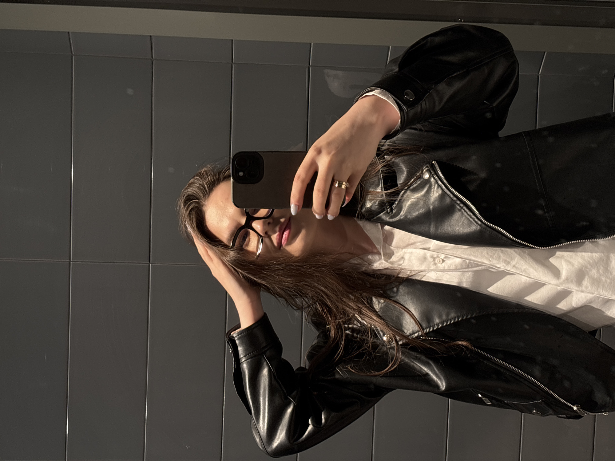 A young woman is captured taking a mirror selfie in an indoor setting. She has long, dark brown hair, wears black-rimmed glasses, a white collared shirt, and a black leather jacket featuring prominent zippers and snap buttons. Her right hand holds a dark-colored smartphone, positioned to take the photo, while her left hand is placed near her temple, gently pushing her hair back. She sports a gold ring on her right ring finger and has light-colored nail polish on her fingernails.
The background consists of dark gray, rectangular wall panels or large tiles, showing subtle reflections and minor surface imperfections. The lighting appears focused, creating highlights on her face, hair, and the textured leather of her jacket, suggesting a specific light source within the room. Her expression is understated, with a slight, contemplative gaze towards the phone. The image location is Izhevsk, Russia.