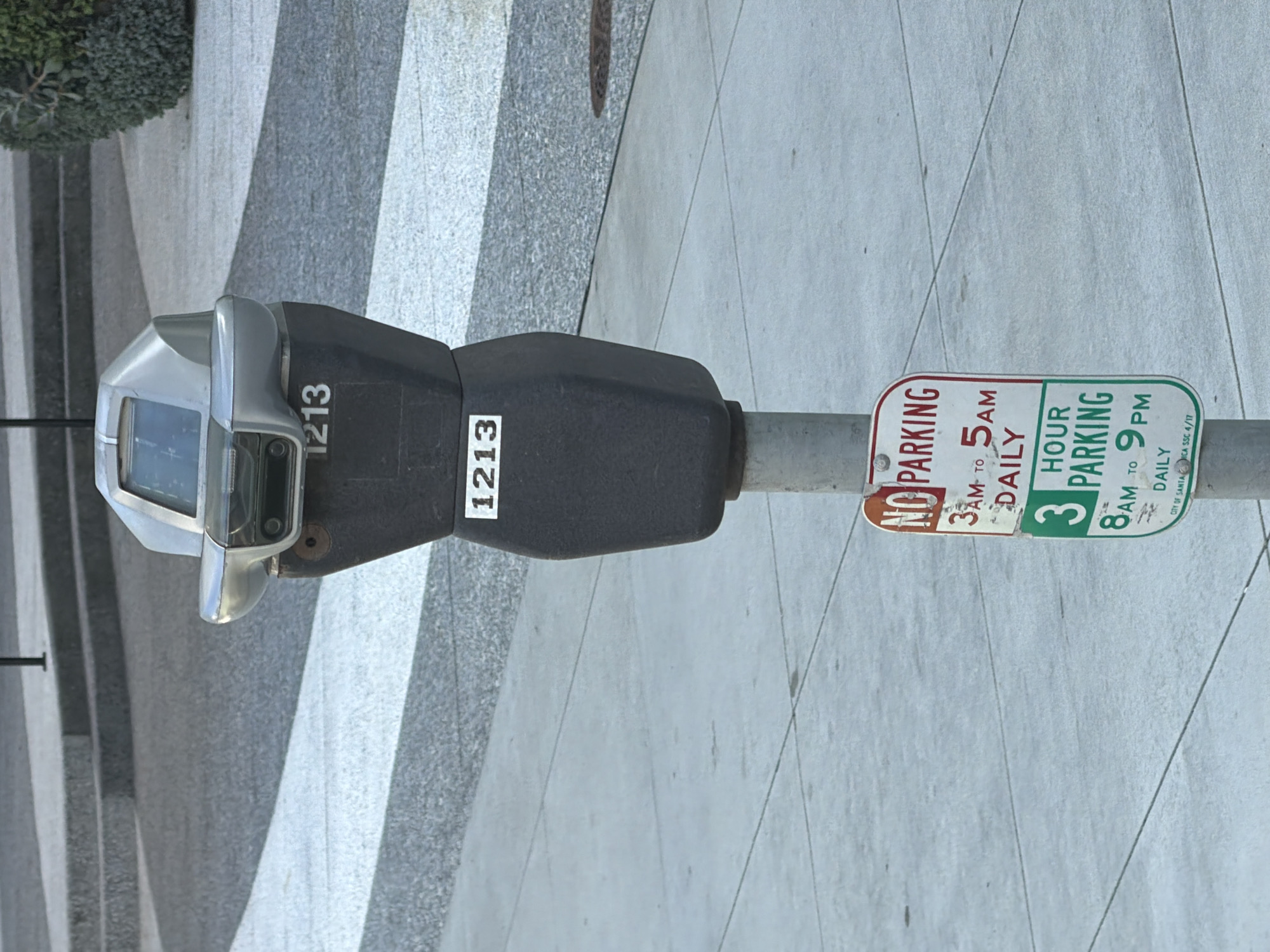 An eye-level, medium shot captures a weathered, dark gray parking meter standing on a concrete sidewalk. The meter has a silver, angular top with a small screen and a black midsection. Stenciled white numbers "1213" are visible on the front of the meter, near the top and also midway down the black section. Below the meter, attached to its metal pole, are two parking restriction signs. The top sign, with a red border, reads "NO PARKING 3 AM TO 5 AM DAILY." The bottom sign, with a green border, reads "3 HOUR PARKING 8 AM TO 9 PM DAILY." At the very bottom of the green sign, small white text indicates "CITY OF SANTA MONICA, CALIF. SCC 4/17." The background shows a diagonal patterned sidewalk with gray and white stripes, and further back, concrete steps and a green shrub. The overall scene is outdoors on a clear day.