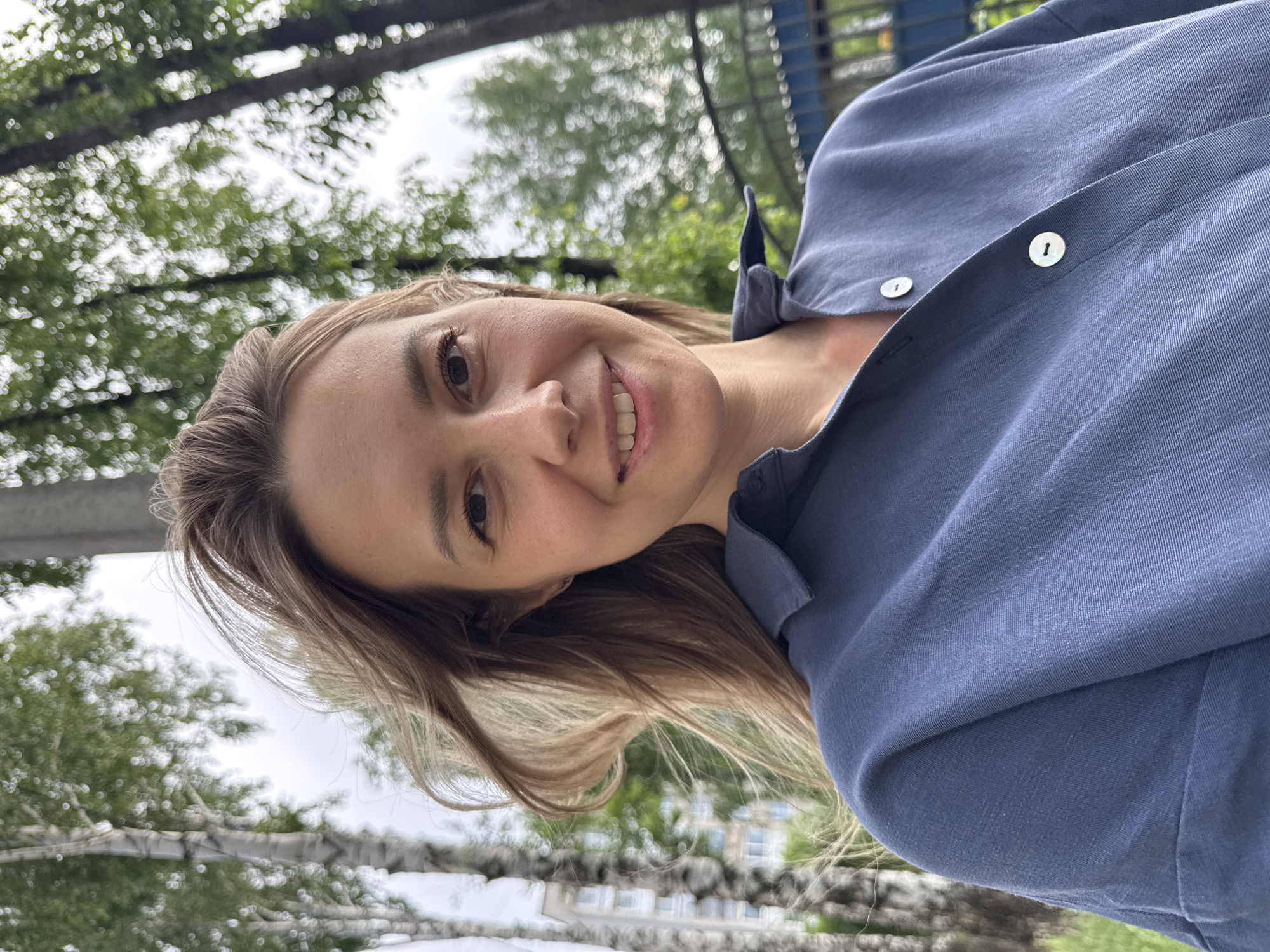 The image is a selfie of a woman with fair skin, smiling at the camera. She has light brown hair and is wearing a blue button-down shirt with white buttons. The background appears to be outdoors, with trees and possibly buildings visible in the distance. Based on the surroundings, the location is likely a park or green space in Chaikovsky, Russia. A fence is visible in the background.