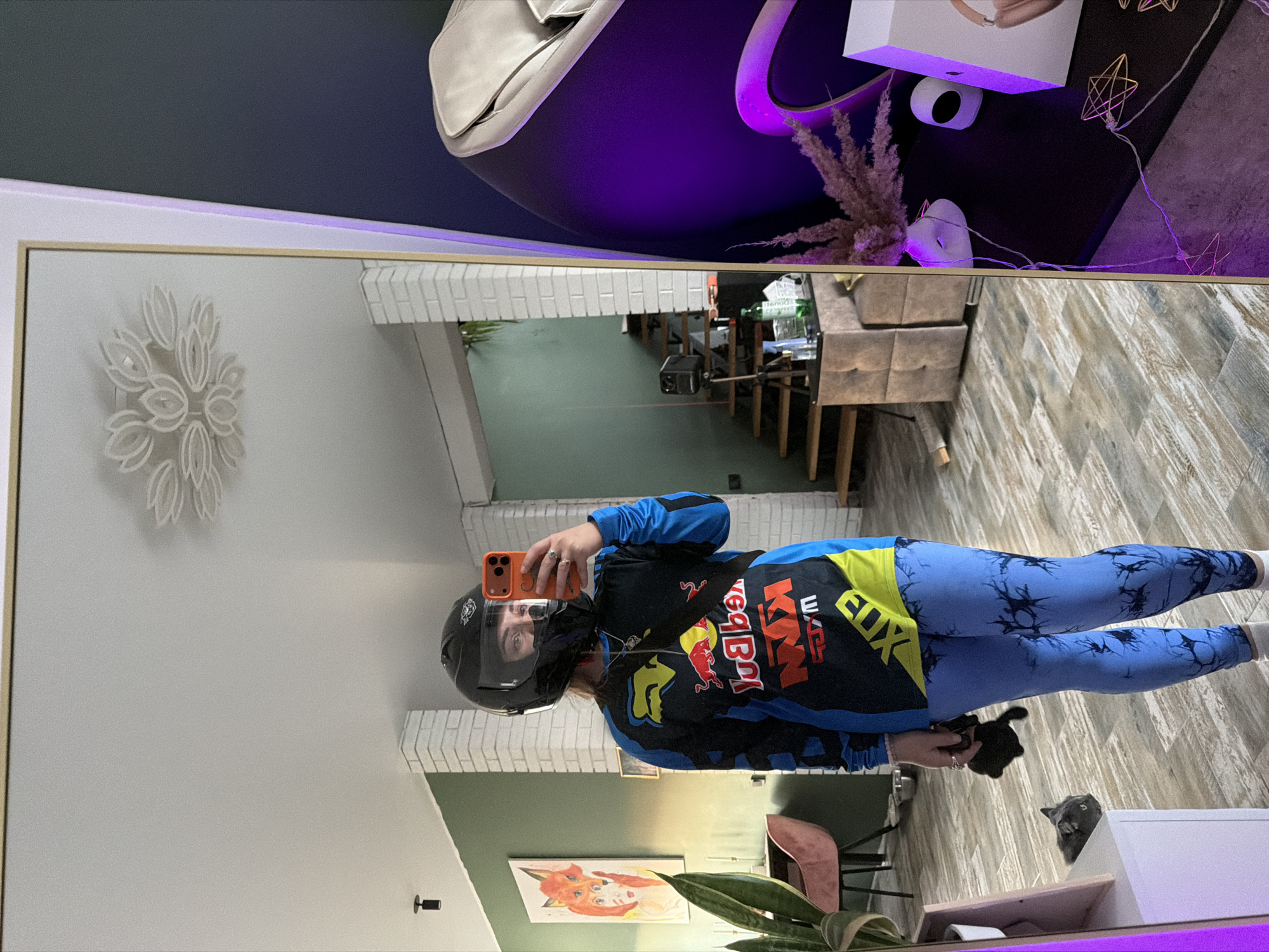 The image shows a reflection in a mirror. A person wearing a motorcycle helmet, a Red Bull KTM jacket, and blue leggings with black tie-dye patterns is taking a selfie with an orange smartphone. The person is indoors, and the reflection reveals a living space with a white brick wall, a decorative wall art piece resembling a flower, a painting of a fox, and a tall green plant. In the background of the reflection is a staircase leading to an upper level and part of a living room. The location is possibly Большая Шильна, Russia. The mirror's reflection also shows a white storage unit with purple lighting, a white object resembling a bag, decorative dried plants, and a section of a room with patterned wallpaper. A black cat is also visible near the base of the mirror.