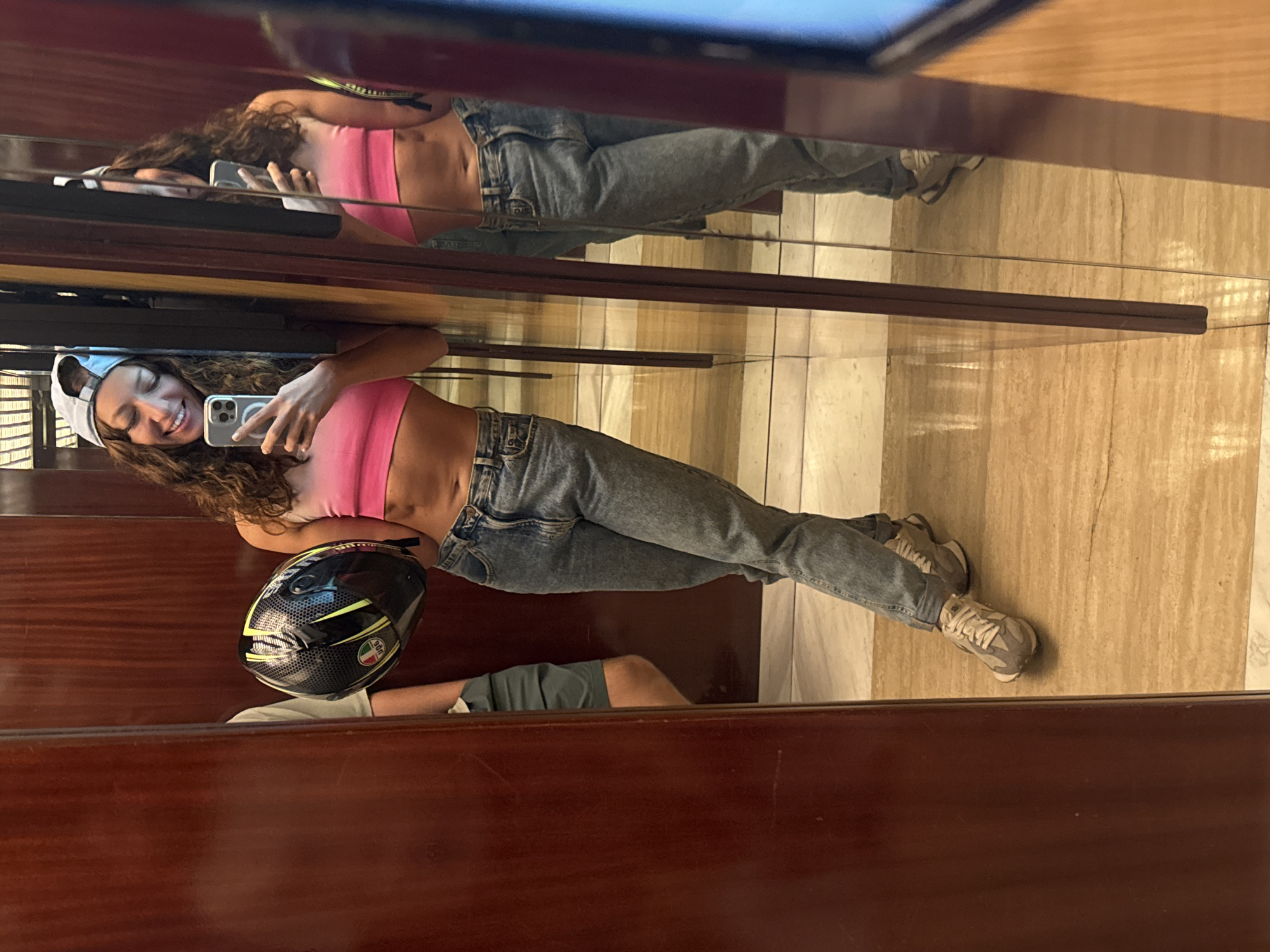 The image, originally captured in a portrait orientation and rotated 90 degrees counter-clockwise, depicts a young woman taking a mirror selfie indoors in Đà Nẵng, Vietnam. The woman, with long, curly brown hair, is wearing a light pink cropped top, light blue jeans, light-colored sneakers, and a light grey baseball cap worn backward. She holds a silver smartphone (likely an iPhone with a triple-lens camera setup) in her left hand, capturing her reflection. A wide, joyful smile illuminates her face, indicating a happy or playful mood.
The setting appears to be an interior space, characterized by dark brown, highly polished wooden paneling on the walls, and a light-colored, possibly marble or tiled floor, visible in the reflection. In the foreground, resting on a wooden surface, is a black motorcycle helmet with striking yellow-green accents and an AGV logo, accompanied by a small Italian flag sticker. A partial reflection of another person's legs, wearing shorts and sneakers, is visible below the main subject, suggesting another individual is present or nearby. The lighting is bright and even, consistent with an indoor environment during daytime or a well-lit evening. The overall scene conveys a casual and modern ambiance.