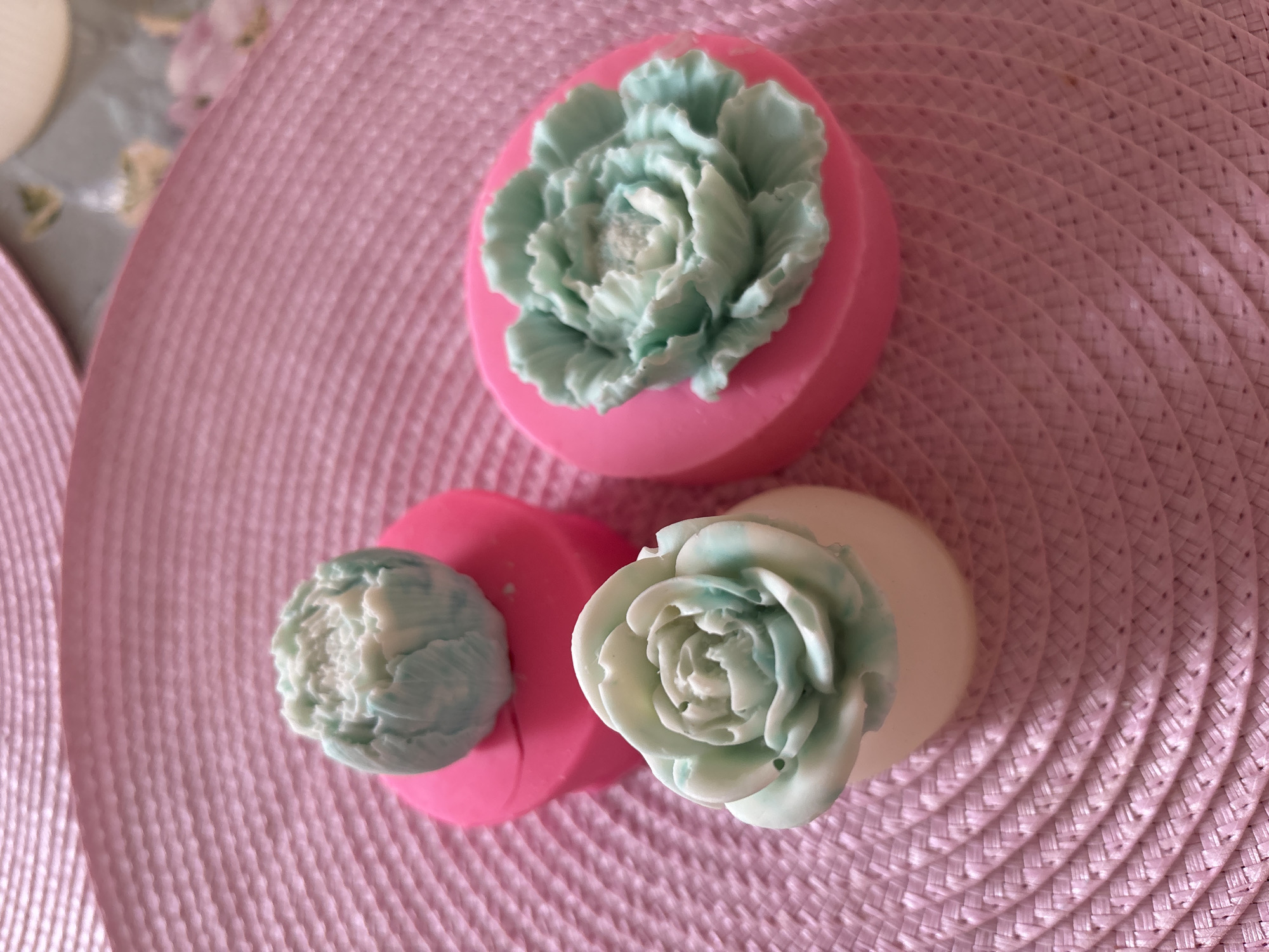 Three decorative soaps, shaped like stylized flowers in shades of pale green and white, are arranged on a pink woven placemat. One soap, a closed bud shape, sits on a bright pink mold. Another, a more open rose shape, sits on a white base and is also on a pink mold. The third soap, a large open flower, rests entirely on a pink mold. The placemat has a textured, circular woven pattern. The background is slightly blurred, suggesting the photo was taken indoors. There is no information available about the location in Naberezhnye Chelny, Russia.