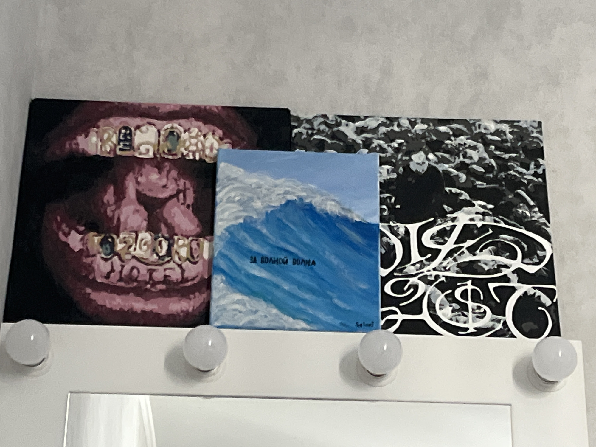 The image shows three small, framed canvases displayed on a wall above a mirror with lights. The leftmost canvas features a close-up painting of pink lips with gold grills, some of which appear to have small gems. The middle canvas is a painting of a large blue wave with white foam, and the text "ЗА ВОЛНОЙ ВОЛНА" is visible on it. The rightmost canvas is a black and white abstract design with white stylized lettering. Below the canvases, the top edge of a mirror is visible, framed by a white surface with several small, round, white lights embedded in it, resembling makeup vanity lights. The overall setting suggests an indoor space, possibly a bedroom or dressing area. There is no visible indication of the city of Chelyabinsk, Russia, or any specific geographical location.