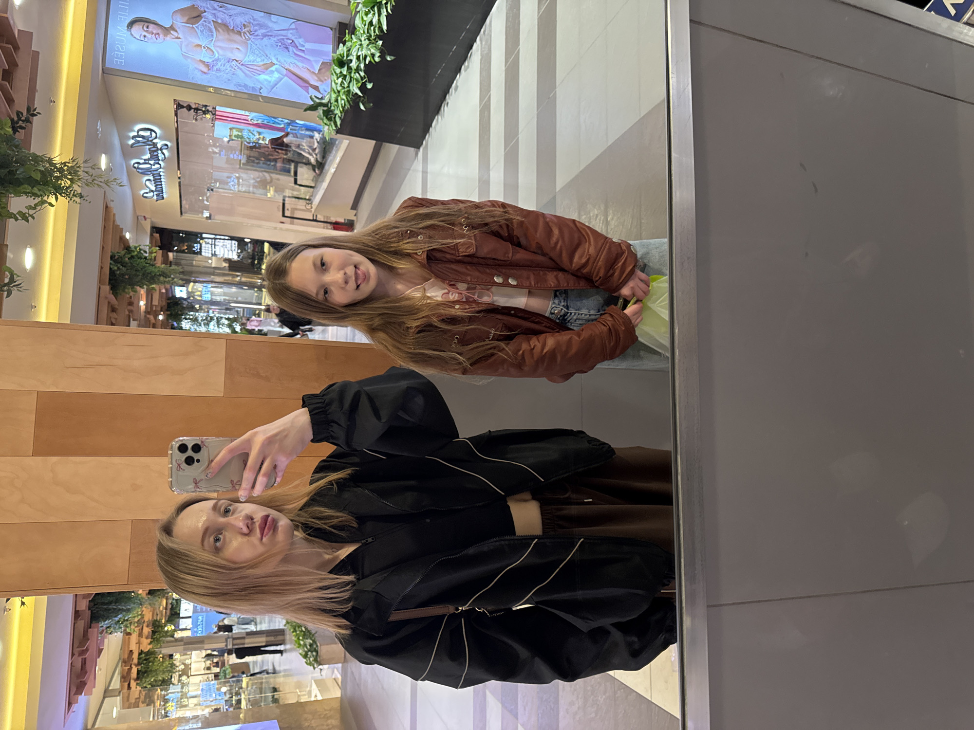 A selfie taken in a mirror shows two women. The woman holding the phone has blonde hair and is wearing a black jacket and brown top. The other woman, reflected in the mirror, has long brown hair and is wearing a brown jacket and jeans. They are standing in a shopping mall, as evidenced by the stores and decorations visible in the background. The exact location in Ol'ginskaya, Russia is not discernible from the image alone.