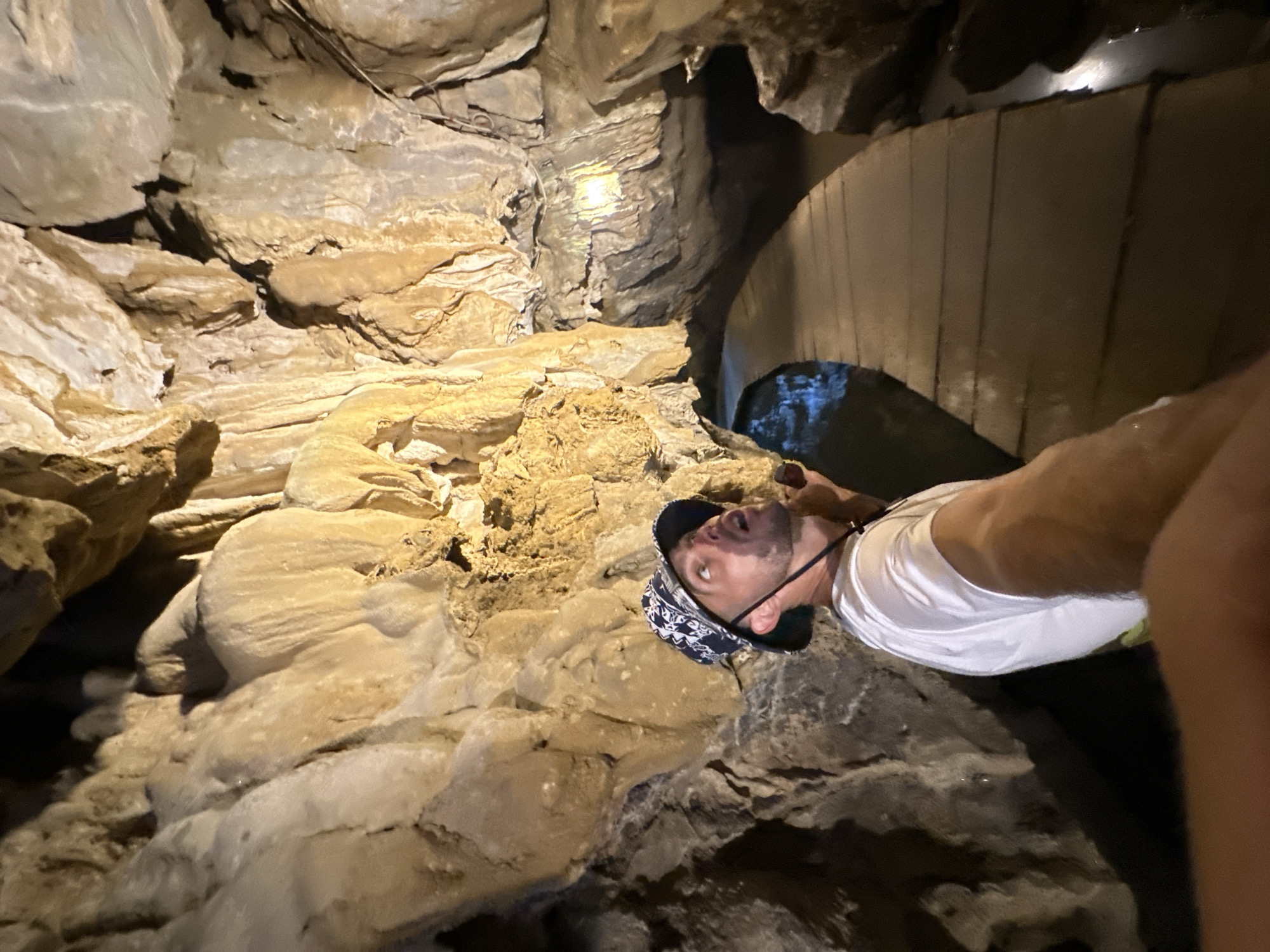 A man wearing a white t-shirt and a patterned bucket hat looks up in awe at the formations inside a cave. The cave walls are rough and textured, with stalactites and stalagmites visible. A wooden walkway is laid out on the cave floor, curving to the right, with dark water visible on either side of the path. The scene appears to be inside Thien Cung Cave on Ha Long Bay in Quảng Ninh province, Vietnam, which is near Yên Sơn Ward.