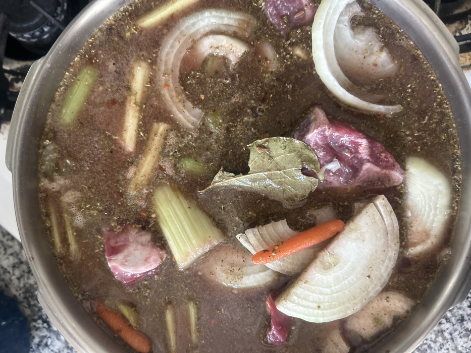 A stainless steel pot is viewed from directly above, containing a dark, reddish-brown liquid base with various raw or partially cooked ingredients simmering. The liquid surface is covered with a fine layer of dried herbs and spices, showing small bubbles indicating heat. Submerged and partially floating within the liquid are irregular pieces of red meat, some exhibiting white fat. Multiple crescent-shaped and ring-shaped slices of white onion are prominently visible. Segments of light green celery stalks and several short, cylindrical orange carrot pieces are also distributed throughout the pot. A single, distinct, ovate, dried bay leaf rests on the liquid surface near the center. The pot sits on an indistinct surface; parts of a light-colored, speckled countertop are visible in the background to the left, and a dark, out-of-focus object, possibly a stovetop grate, is visible to the right. No individuals are present. The specific location within Los Angeles, United States of America, cannot be determined from the visual evidence.