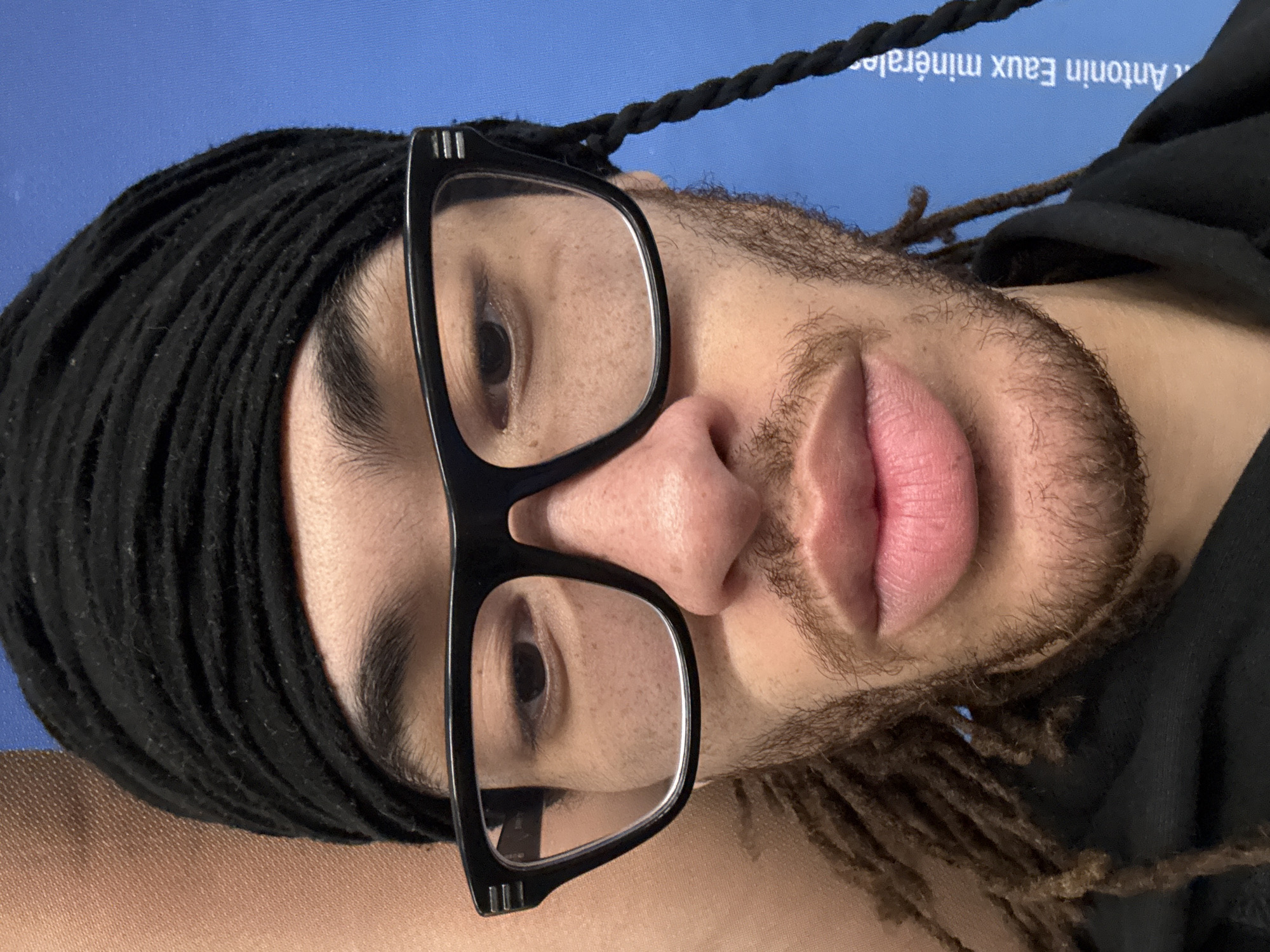 A close-up portrait shows a person with light brown skin, dark brown eyes, and a short beard. They are wearing black-framed glasses and a black head covering. Some of their hair, styled in dreadlocks or braids, is visible. In the background, indistinct text including "Antonin Eaux minérales" suggests the image may have been taken in Paris, France, as this brand of mineral water is available there. The image appears to have been rotated.