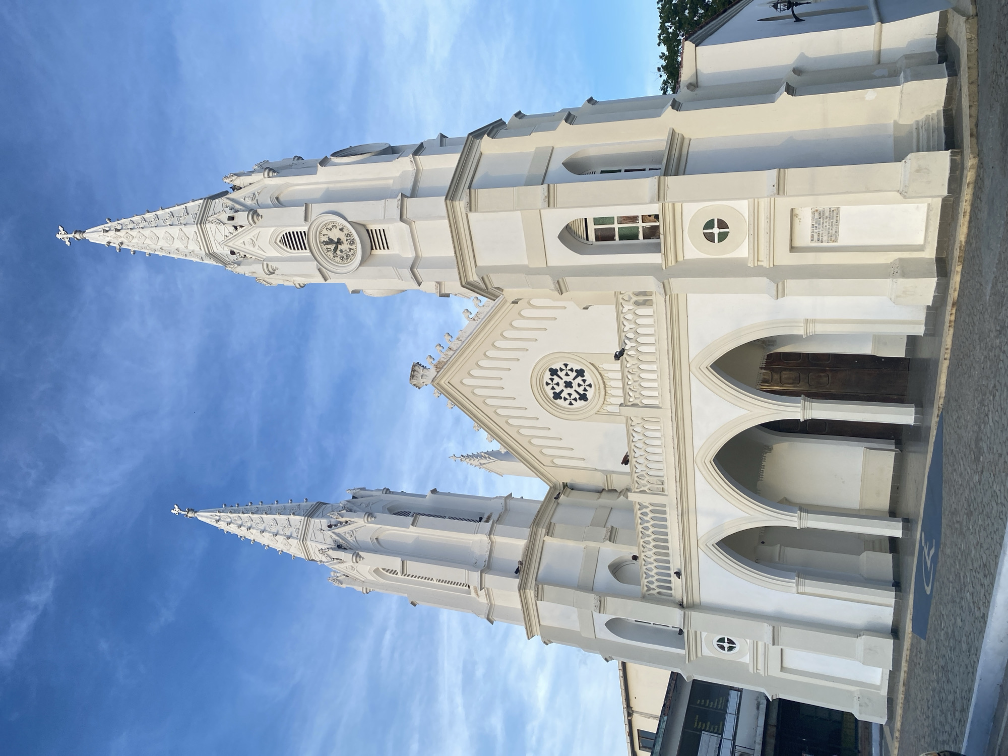 The image depicts the Iglesia de San Juan Evangelista, a historic white church located in Juan Griego, Venezuela, set against a bright blue sky with streaky clouds. The church exhibits a gothic architectural style, characterized by its pointed spires, arched windows, and intricate details.
The church has two prominent spires reaching high into the sky. A clock face is visible on the higher spire. The main section of the church features a series of arched entryways leading to a set of doors. Architectural details such as circular windows and ornate railings adorn the building. A plaque is noticeable on the church's facade. The foreground shows part of the street and a glimpse of a modern building adjacent to the church.