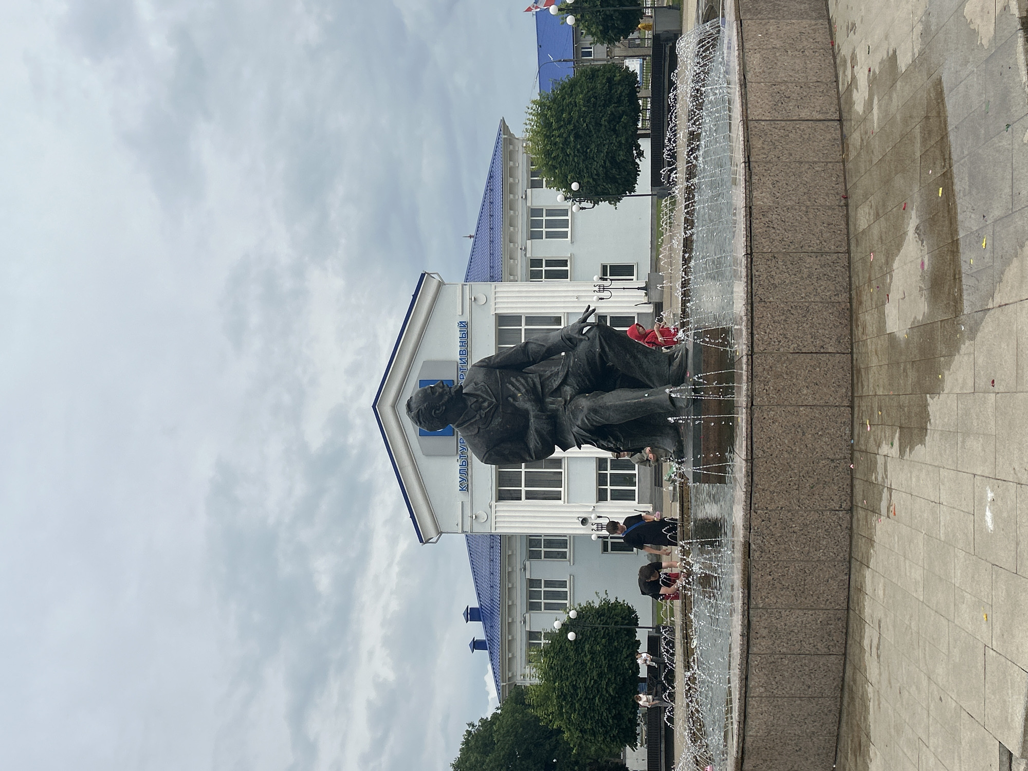 In the city of Chaikovsky, Russia, a bronze statue of a seated man, presumably Pyotr Ilyich Tchaikovsky, is the central focus of a public square. The statue sits in front of a light blue building with a blue roof, which appears to be a cultural center or similar establishment. The building has white columns and large windows. A fountain surrounds the base of the statue, with water spraying in a wide arc. Several people are visible around the fountain and in front of the building, some interacting with the water. The ground is paved with stone tiles, some of which are wet from the fountain. Trees line the sides of the square, and the sky is overcast.