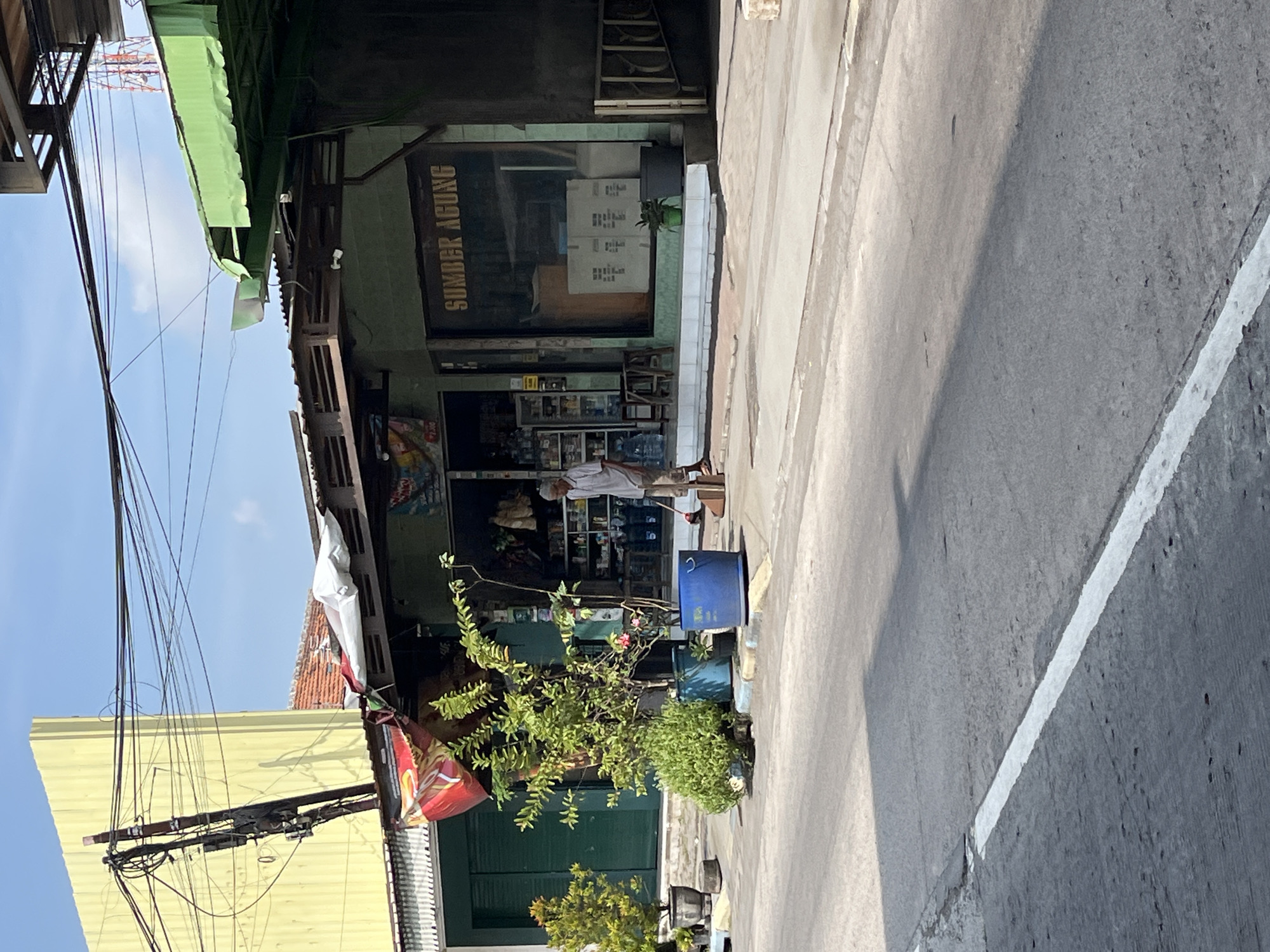 The image shows a street scene in Boro, East Java. In the foreground, a gray asphalt road with a white dashed line is visible. A shadow of a building falls across the road. On the right side of the road, a sidewalk is lined with various potted plants. Behind the sidewalk, a green-fronted shop with the text "SUMBER AGUNG" in gold lettering is present. The shop has large glass windows and displays shelves of goods inside. An elderly man in a light-colored shirt and pants is sweeping the sidewalk in front of the shop with a broom. To the left of the shop, a yellow building with a corrugated metal roof and a prominent utility pole with numerous wires is situated. There are also green metal roll-up doors on the building. The sky is clear with some clouds.