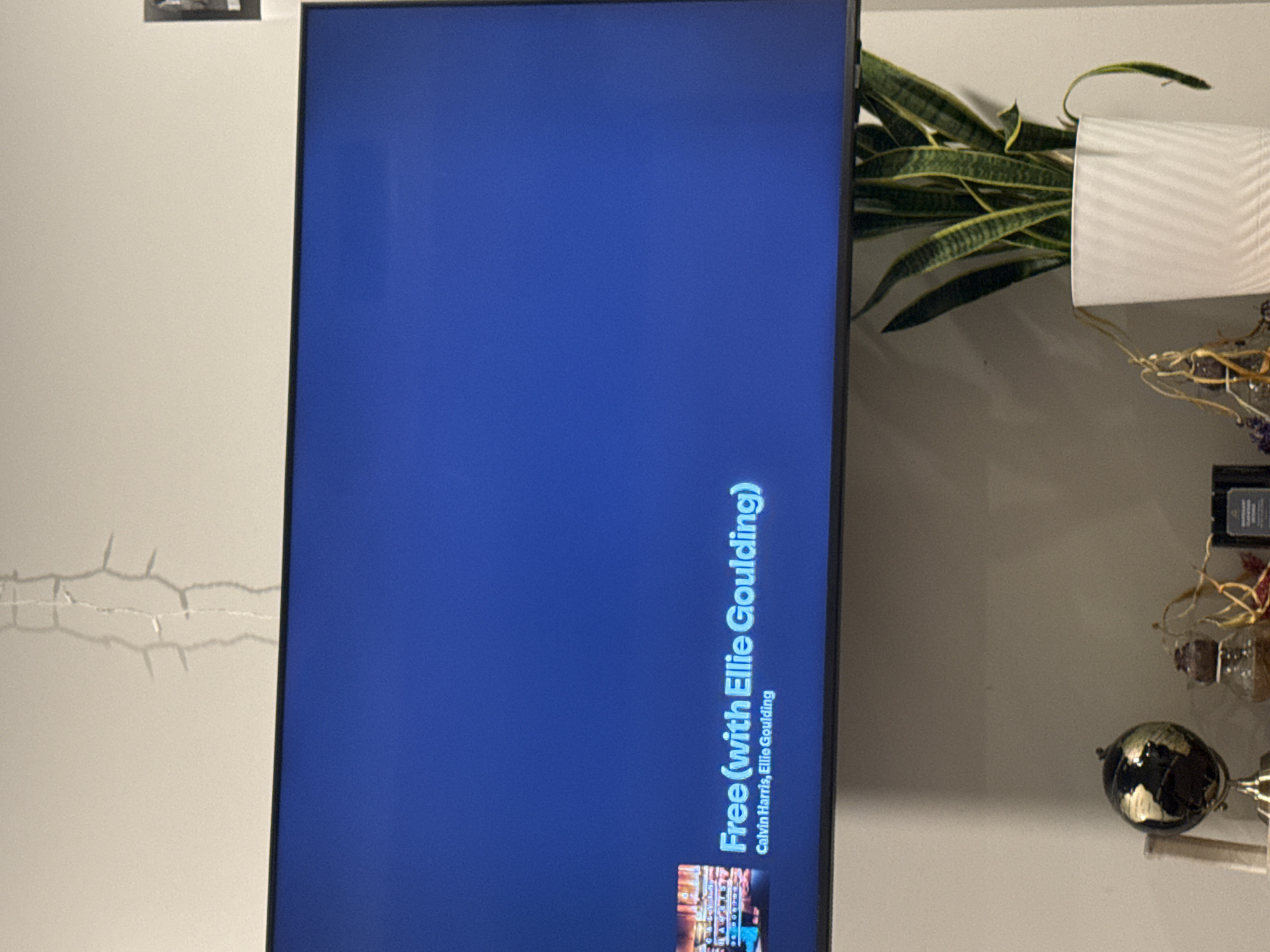 The image shows a close-up of a television screen displaying the song "Free (with Ellie Goulding)" by Calvin Harris. To the right of the TV is a plant in a white pot. Below the plant is a small globe and other decorative items. To the left of the TV, the wall has visible cracks. The location is likely a residential interior, but there is no information to determine if the location is in Austin, United States of America.