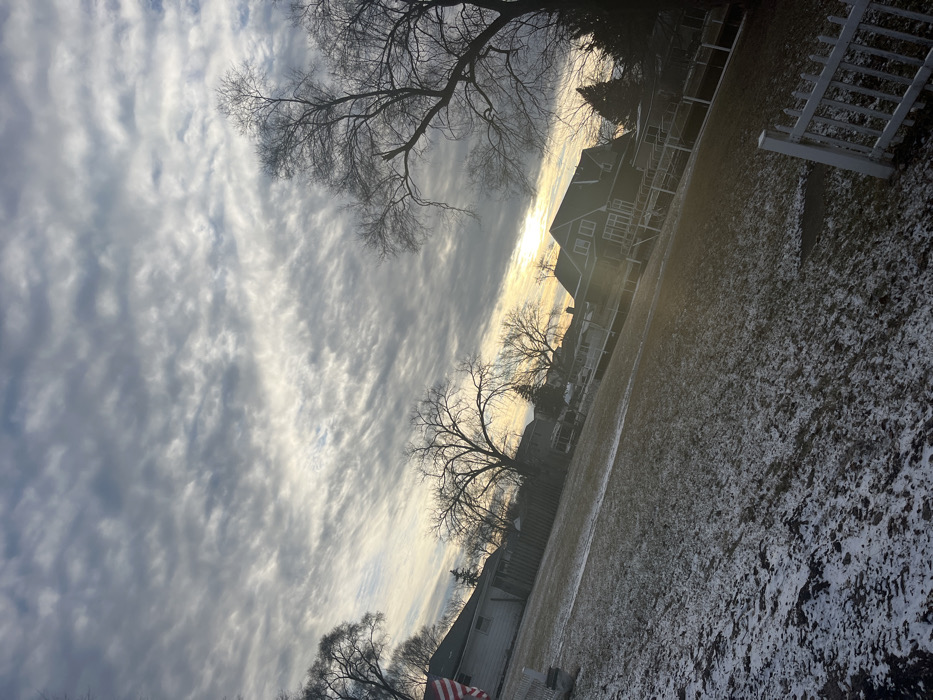 An angled view of a winter scene shows a sky filled with streaky clouds, partially obscuring the sun. Bare trees frame the scene. Below, a snowy yard extends towards a fence and houses. The light from the sun creates a bright glow behind the houses, highlighting the barren branches of the trees.