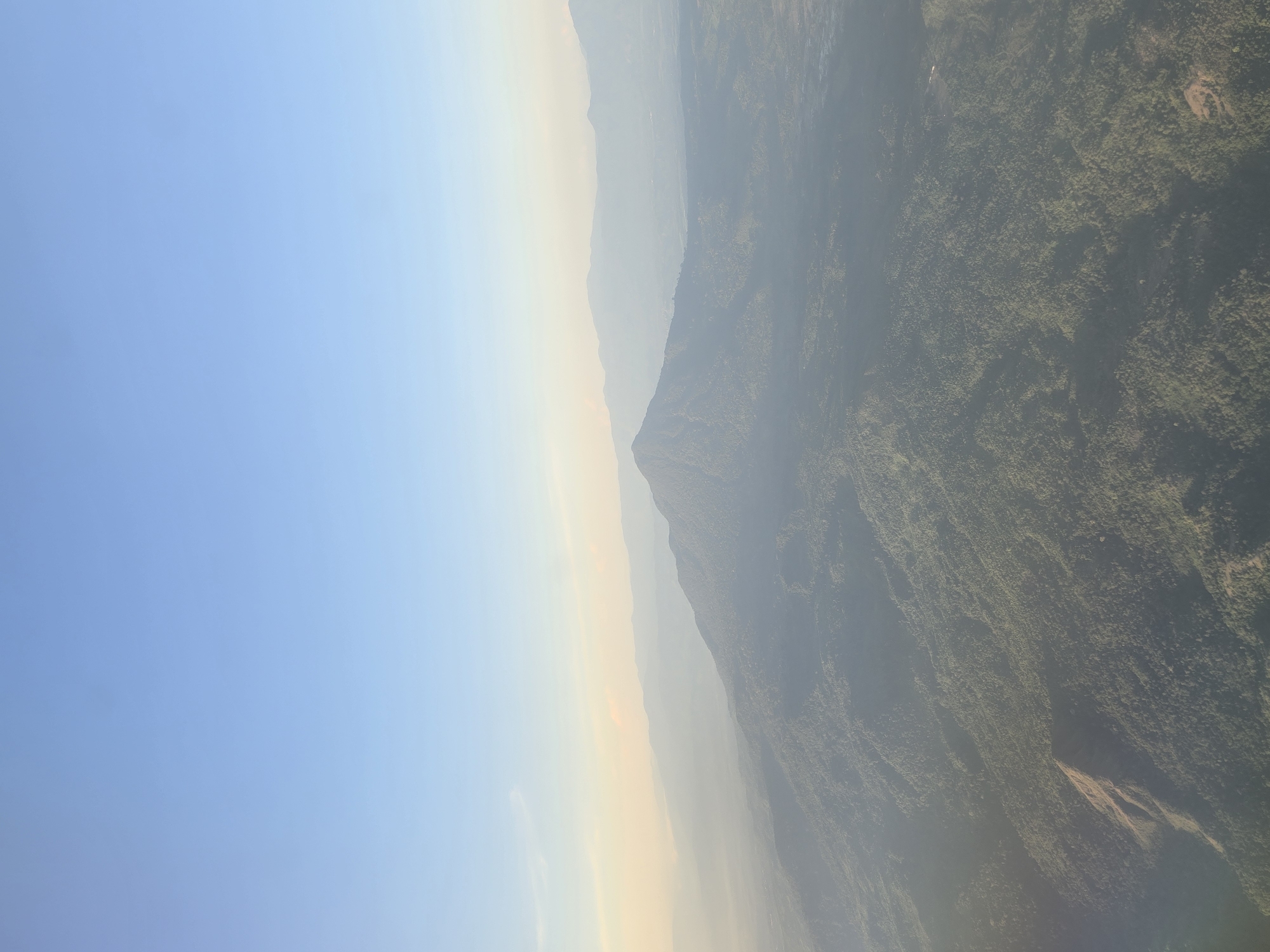 An aerial view captures a majestic, cone-shaped mountain peak rising from a vast, verdant landscape. The mountain is covered in dense green foliage, with a prominent ridge extending from its base. The surrounding terrain is also densely forested, with rolling hills and valleys stretching towards the horizon. In the distance, a hazy layer of mist softens the outline of more distant mountain ranges. The sky above is a clear, pale blue, with a faint band of warmer, lighter hues near the horizon, suggesting either sunrise or sunset. The overall scene is one of natural beauty and vastness. The location is Ujongkeupula, Indonesia.