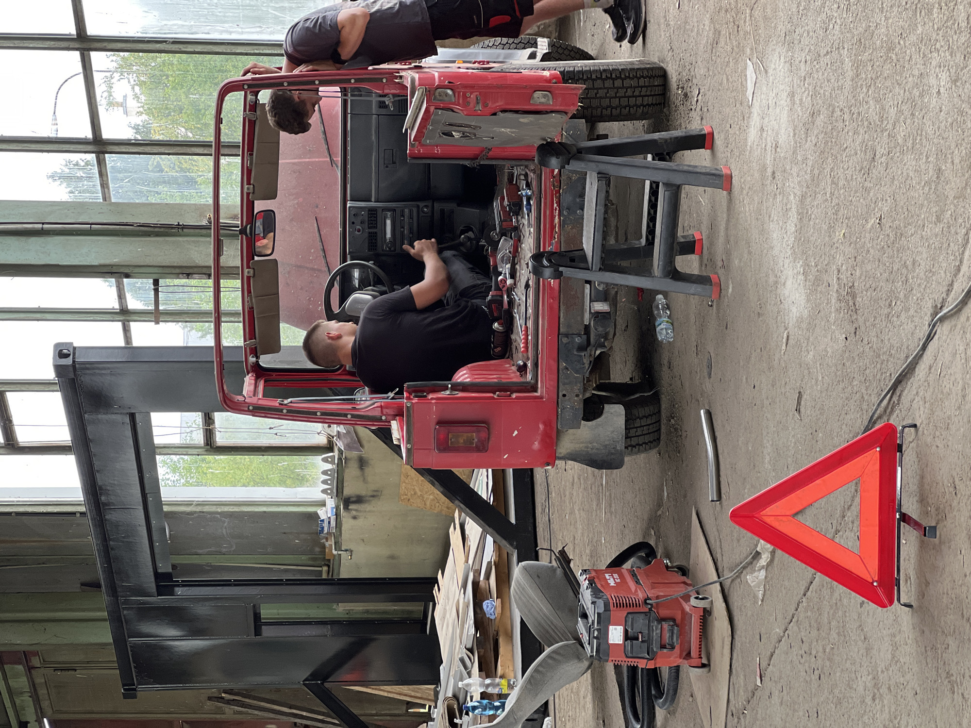 In an industrial workshop located in Lanžhot, Czechia, two men are working on a red Jeep Wrangler that has been partially disassembled. The vehicle is positioned centrally in the frame, with its doors and roof removed. One man, wearing a black t-shirt, is seated in the driver's seat, appearing to be operating the gear stick. The other man, in a grey t-shirt, is standing outside the vehicle, leaning in towards the dashboard, possibly inspecting or working on components.
The workshop environment is visible with large windows in the background, allowing natural light to enter, illuminating the dusty concrete floor and scattered tools and materials. To the left of the Jeep, a car seat is placed on the floor, alongside a red HILTI vacuum cleaner. A red triangle-shaped warning sign is prominently displayed in the foreground, resting on the concrete floor. The overall scene suggests an active repair or restoration project within a functional but cluttered workspace.
