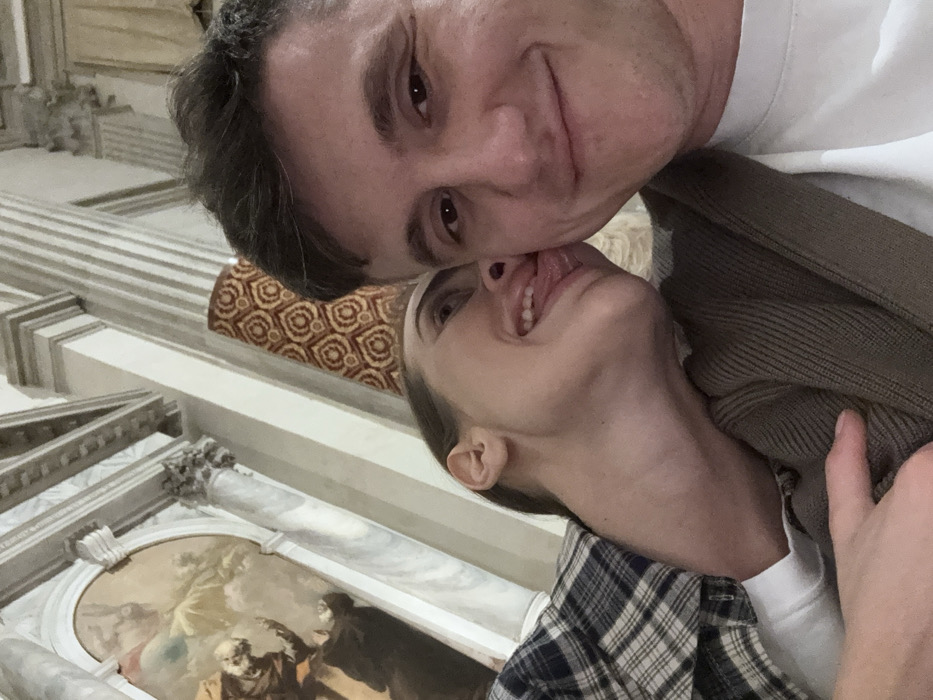 A close-up, upward-angled selfie photograph depicts a male and a female in the interior of the Scuola Grande di San Marco in Venice, Italy. The male, positioned on the right and facing the camera, has short dark hair and a faint smile. He wears a white t-shirt under a brown ribbed cardigan. The female, positioned to the left and slightly behind the male, has her brown hair pulled back. She wears a blue and white plaid shirt over a white t-shirt and has her mouth open, extending her tongue towards the male's right cheek. Her left hand rests on the male's left arm. The background features classical architectural elements, including fluted marble columns and elaborate cornices, typical of the historic Venetian building. On the far left, an arched section of a large religious oil painting, identifiable as Tintoretto's "The Miracle of St. Mark Freeing the Slave," is visible. A patterned reddish-brown and beige textile hangs vertically in the upper center-right background.