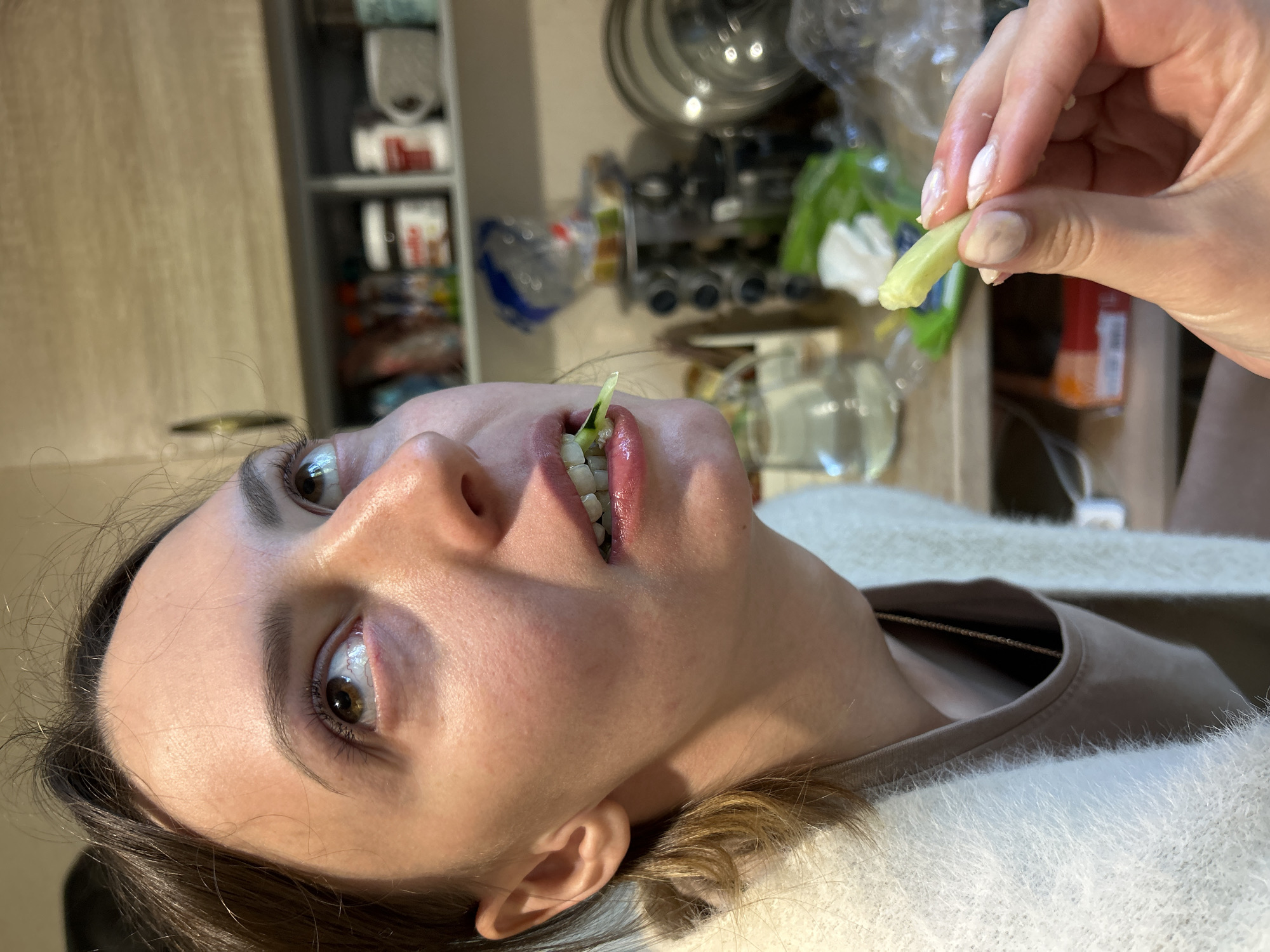 In this close-up, vertically oriented image, a person is having a piece of green vegetable, possibly a spring onion or a sliver of cucumber, placed into their mouth. The person's mouth is open, revealing their teeth, and the vegetable is partially inserted. Their eyes are open, looking upwards, with a neutral or slightly surprised expression. The person has light brown hair and fair skin. They are wearing a light-colored, fuzzy sweater.
The background appears to be a kitchen or pantry area. Shelves filled with various items, including what looks like food packaging and containers, are visible. A metal pot or pan is also in the background, suggesting a domestic setting. The lighting is diffused and appears to be from an indoor source, indicating it is likely daytime, but not necessarily bright daylight.
The person's right hand, with neatly manicured nails, is visible at the top right of the frame, holding the vegetable. The focus is primarily on the person's face and the interaction with the food item. No text is discernible in the image. The scene is set in Khimki, Russia.