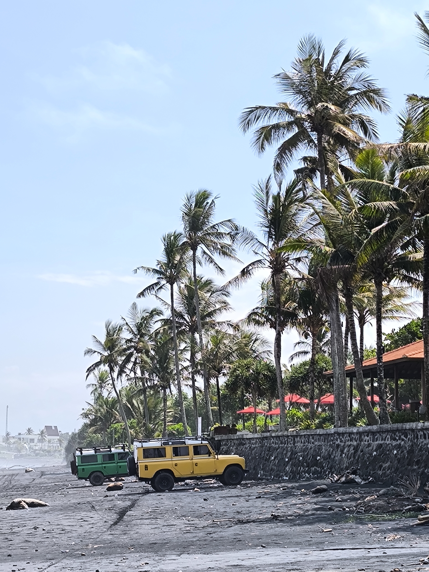 The image shows a beach scene in Selemadeg Timur, Indonesia. Two vintage Land Rover vehicles, one green and one yellow, are parked on a black sand beach. A row of palm trees lines the back of the beach, along with a low stone wall and a building with a red tile roof. In the background, there is a structure resembling a tower and several buildings. The sky is a pale blue with some clouds. Debris is scattered on the beach, particularly near the stone wall.