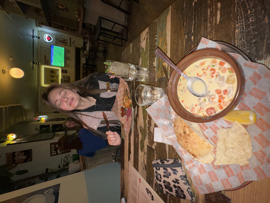 An interior shot depicts a casual restaurant or bar, identified as Murzik Brazik in Moscow, Russia, during what appears to be evening hours. A young adult female, with long brown hair, is seated at a rustic wooden table in the foreground, eyes closed and exhibiting a subtle smile. She holds a knife in her right hand and a fork in her left, poised over a plate containing cooked food, possibly meat, served on branded paper with a side of red sauce and pickled vegetables.
On the table in front of her are two glasses of water; one contains ice, a lemon slice, and mint, suggesting a beverage. A leopard-print wallet or clutch and a menu are visible to her left. In the lower frame, a ceramic bowl holds a creamy soup with red peppers, mushrooms, and other ingredients, with a spoon resting within it. This bowl is placed on a metal tray lined with patterned paper, featuring the text "МУРЗИК БРАЗИК". Two pieces of flatbread and a lemon wedge accompany the soup.
The background reveals dim, atmospheric lighting. The walls exhibit a worn, industrial aesthetic, adorned with various framed pictures, including a "BEER" poster, a four-figure image resembling The Beatles' "Abbey Road" album cover, and several neon signs. A flat-screen television mounted on the wall displays a soccer match. Other patrons are visible in the blurred background, seated at tables with high stools.