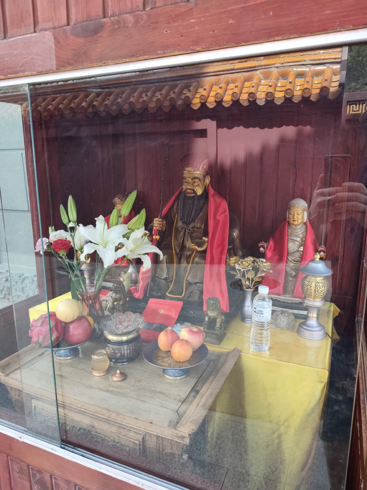 A glass-enclosed display case, likely situated in Daqing, China, contains religious statuary and offerings. In the center of the display, two figures are prominently featured. The larger figure on the left is a man with a dark beard, wearing a red scarf draped over his shoulders. To his right stands a smaller, seated figure in similar attire. In front of the statues, a wooden table holds various items. A vase of white lilies and red carnations sits on the left, next to a collection of fruits including dragon fruit, peaches, and a yellow melon. Further to the right, an incense burner with lit incense sticks is visible, along with a small golden jar and a bottle of water. A decorative lamp with intricate carvings is positioned on the right side of the table. The background of the display is a dark wooden paneling. The entire scene is viewed through glass, with reflections visible on its surface.