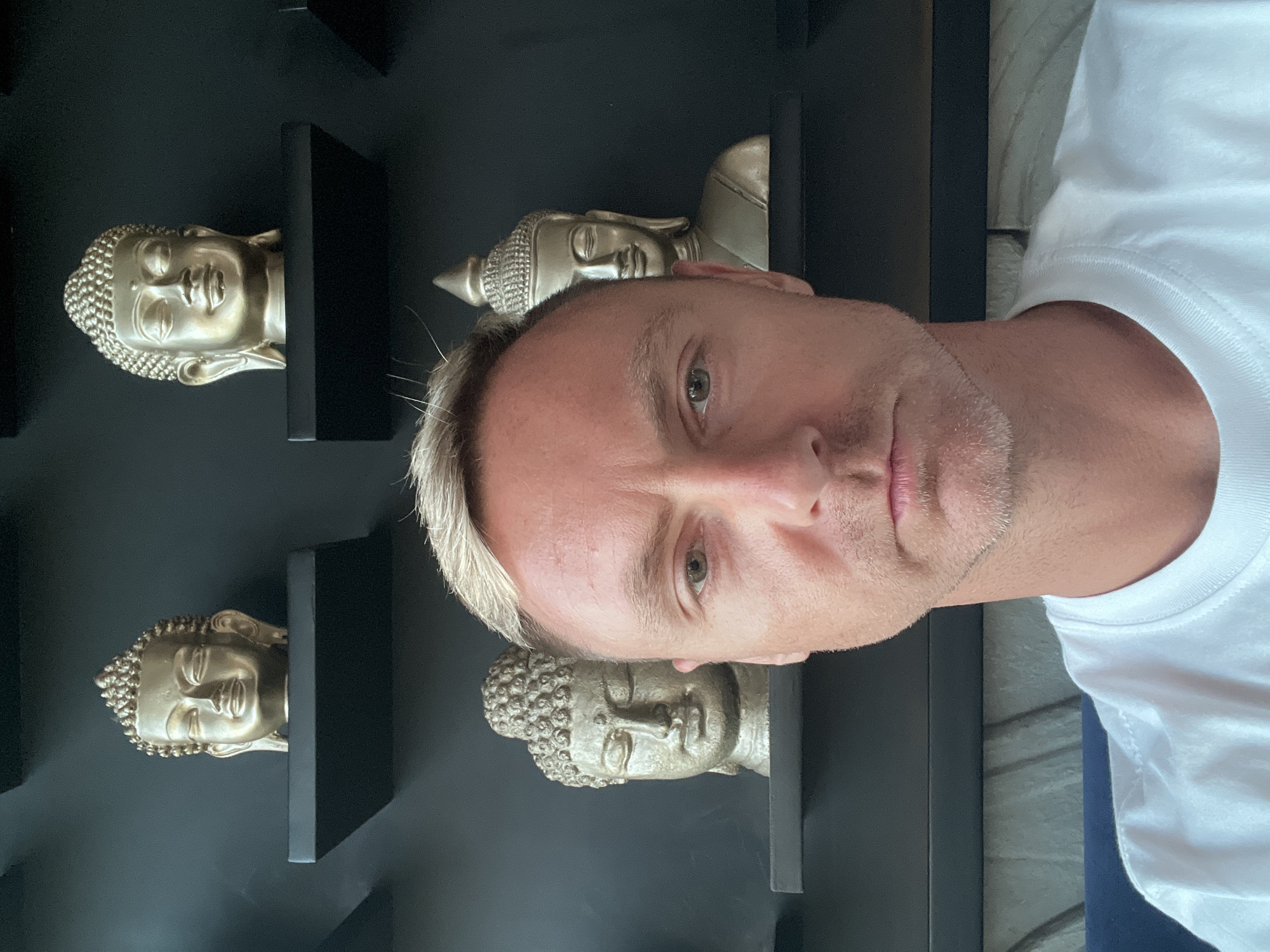 A medium shot selfie of a fair-skinned man with blond hair and a stubbled jawline is taken in front of a dark wall with several gold-toned Buddha busts placed on black shelves. The man is wearing a white t-shirt and looking directly at the camera with a neutral expression. The Buddha busts are of varying sizes and designs, with some appearing to be full heads and others depicting the upper torso. The shelves are arranged in a staggered pattern, with some placed higher and others lower on the wall. The background is dark and out of focus, with subtle lighting highlighting the Buddha busts. The overall scene appears to be indoors, possibly in a home or a decorative space.