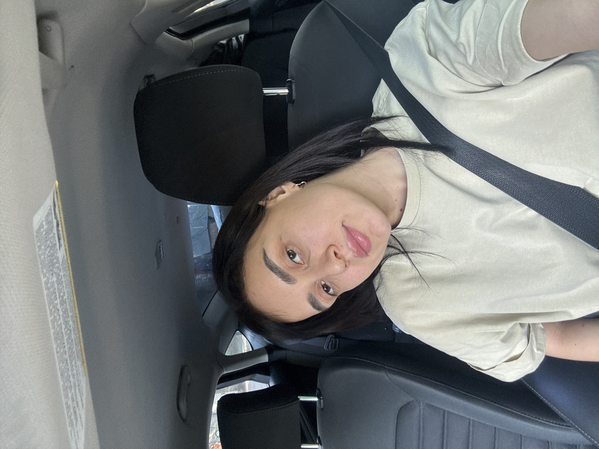 A young adult female is seated in the passenger seat of a car, wearing a seatbelt and a light-colored t-shirt. She has dark hair and is facing the camera. The interior of the car is visible, including the headrest and seat. The location is likely Tbilisi, Georgia, but there are no visual cues within the image to confirm this.