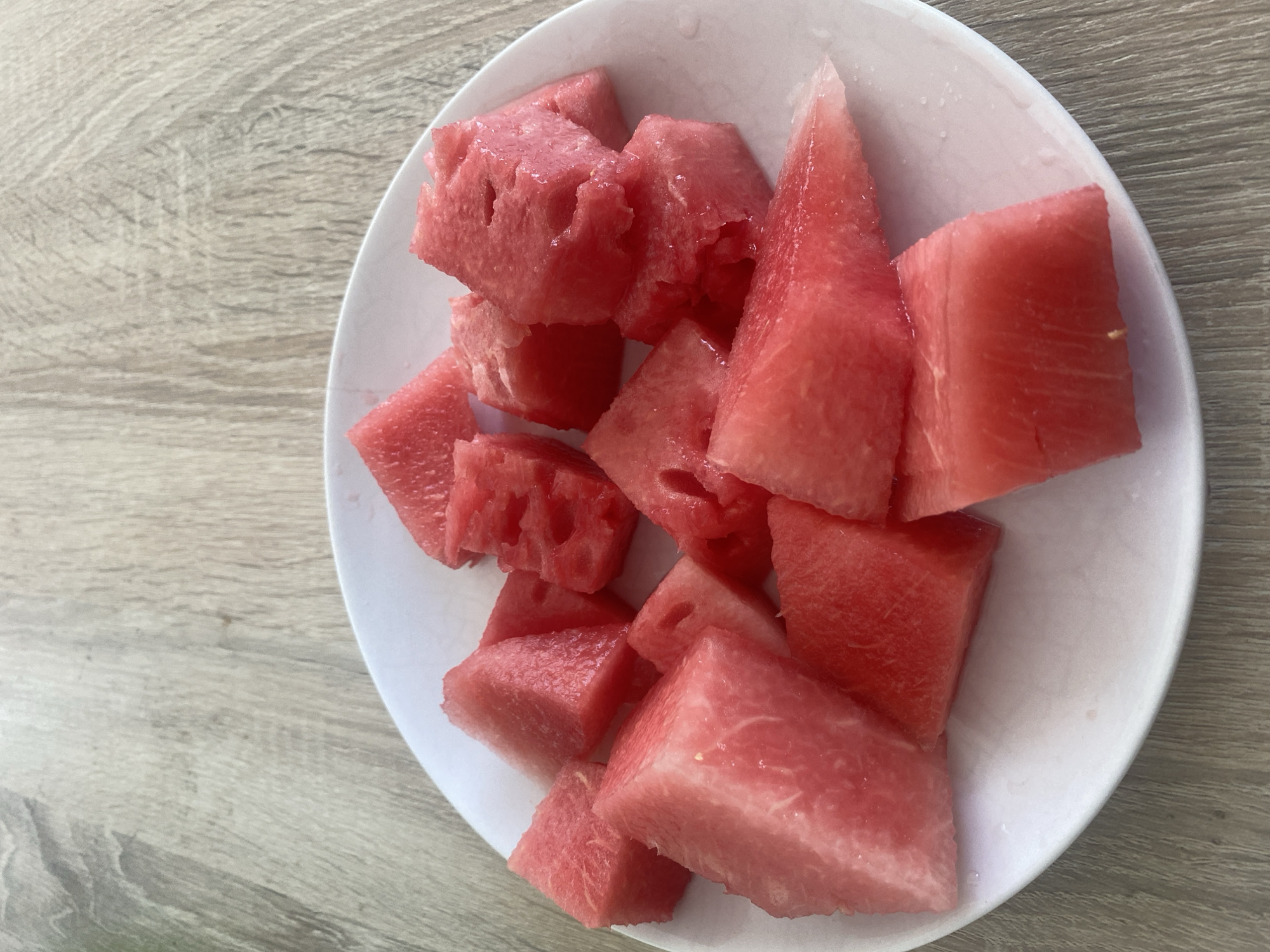A plate of cut watermelon sits on a wooden table. The watermelon pieces are various sizes and shapes, some triangular, some cubic. The plate is white and circular. The wooden table has a light brown, wood-grain pattern. There are no people or animals visible in the image. The location is unknown, but the food suggests a warm climate.