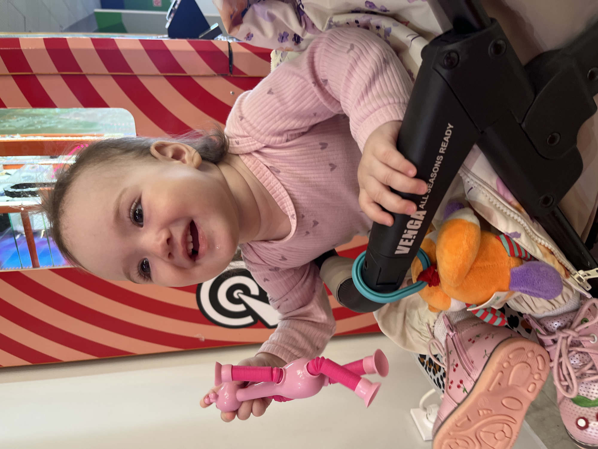 A baby in a pink onesie is sitting in a stroller. The baby is holding a pink toy in her left hand and the stroller handlebar in her right. Inside the stroller is a stuffed animal and a pair of pink shoes. The stroller is next to a red and white striped structure. The location is Shushary, Russia.