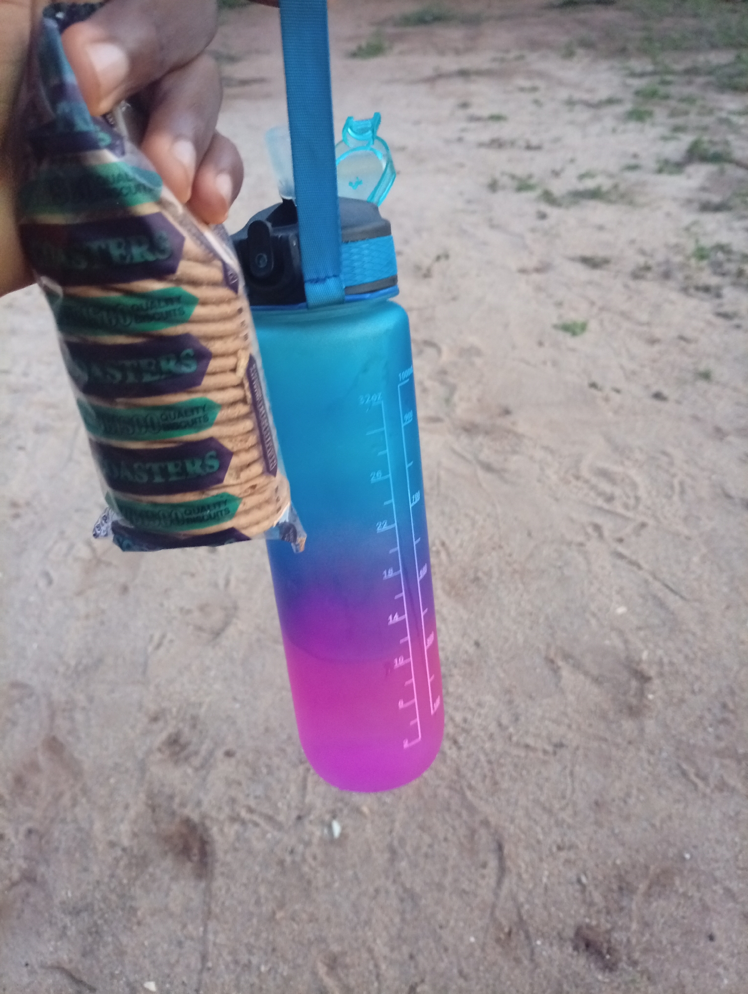 A hand holds a package of Roasters brand sweet gourmet biscuits next to a blue and purple ombre water bottle with white measurement markings. The background consists of a sandy ground with some sparse greenery. This image was likely taken in Ikorodu, Nigeria.