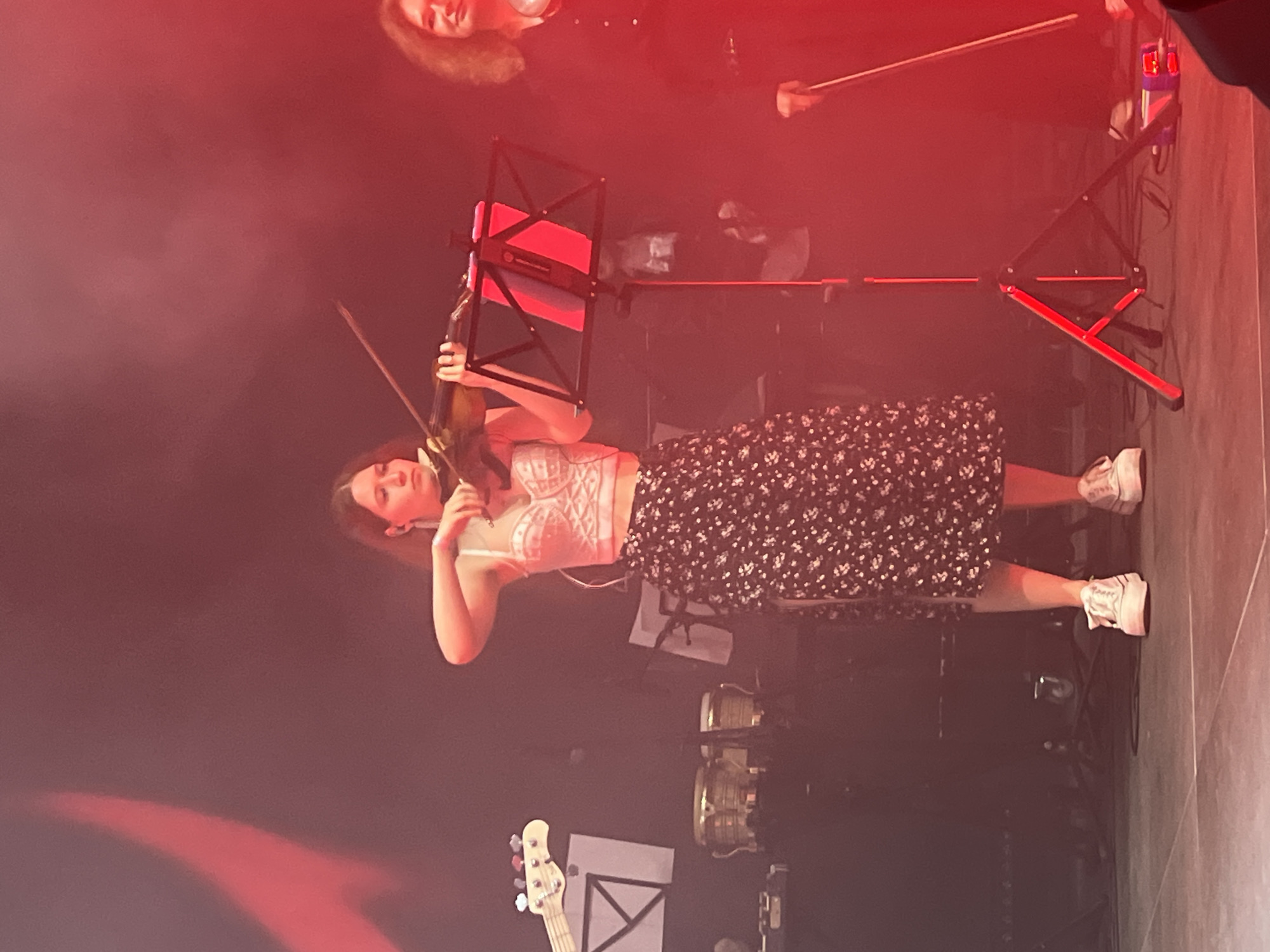 A young woman stands on a stage, illuminated by red stage lights, holding and playing a violin. She wears a white, ornate top and a dark, floral-patterned skirt, along with white sneakers. A music stand with red accents is positioned in front of her. In the background, other musicians and instruments, including a bass guitar and percussion instruments, are faintly visible. The scene is set on a stage, likely in a performance venue. While it is not possible to determine the specific location in Saint Petersburg, Russia, from the given image, the event is likely a musical performance in a city setting.