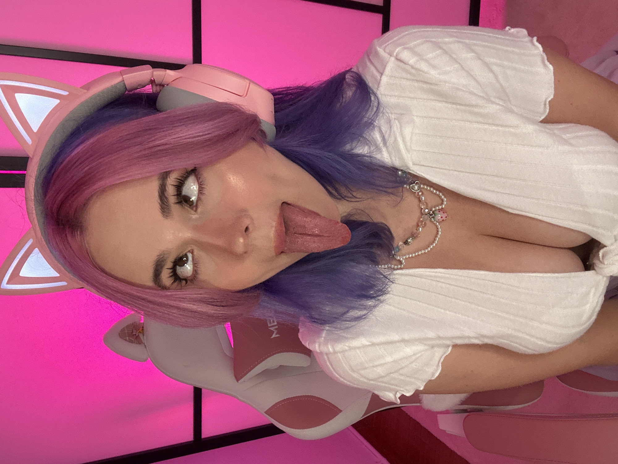 A young woman with pink and purple hair is sitting in a pink and white gaming chair. She is wearing a white, ribbed short-sleeved shirt and a pearl necklace with a strawberry charm. She has pink cat-ear headphones on and is sticking her tongue out, looking up. The background is a bright pink wall with a grid pattern. This image does not contain any recognizable landmarks or features that would allow for its location to be identified as Oviedo/uviéu, Spain.
