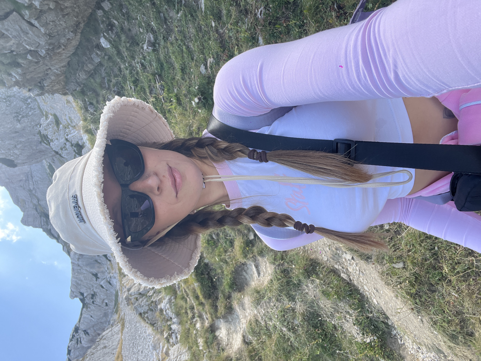 A young woman with light brown hair styled in two braids, wearing a bucket hat, sunglasses, a white crop top with pink trim, and pink long-sleeved shirt is taking a selfie while hiking in a mountainous area near Боричје, Montenegro. The background shows rocky terrain and a steep, winding path, suggesting an outdoor adventure in a natural, rugged environment. The woman's attire indicates preparation for an outdoor activity like hiking. The lighting suggests it is daytime.