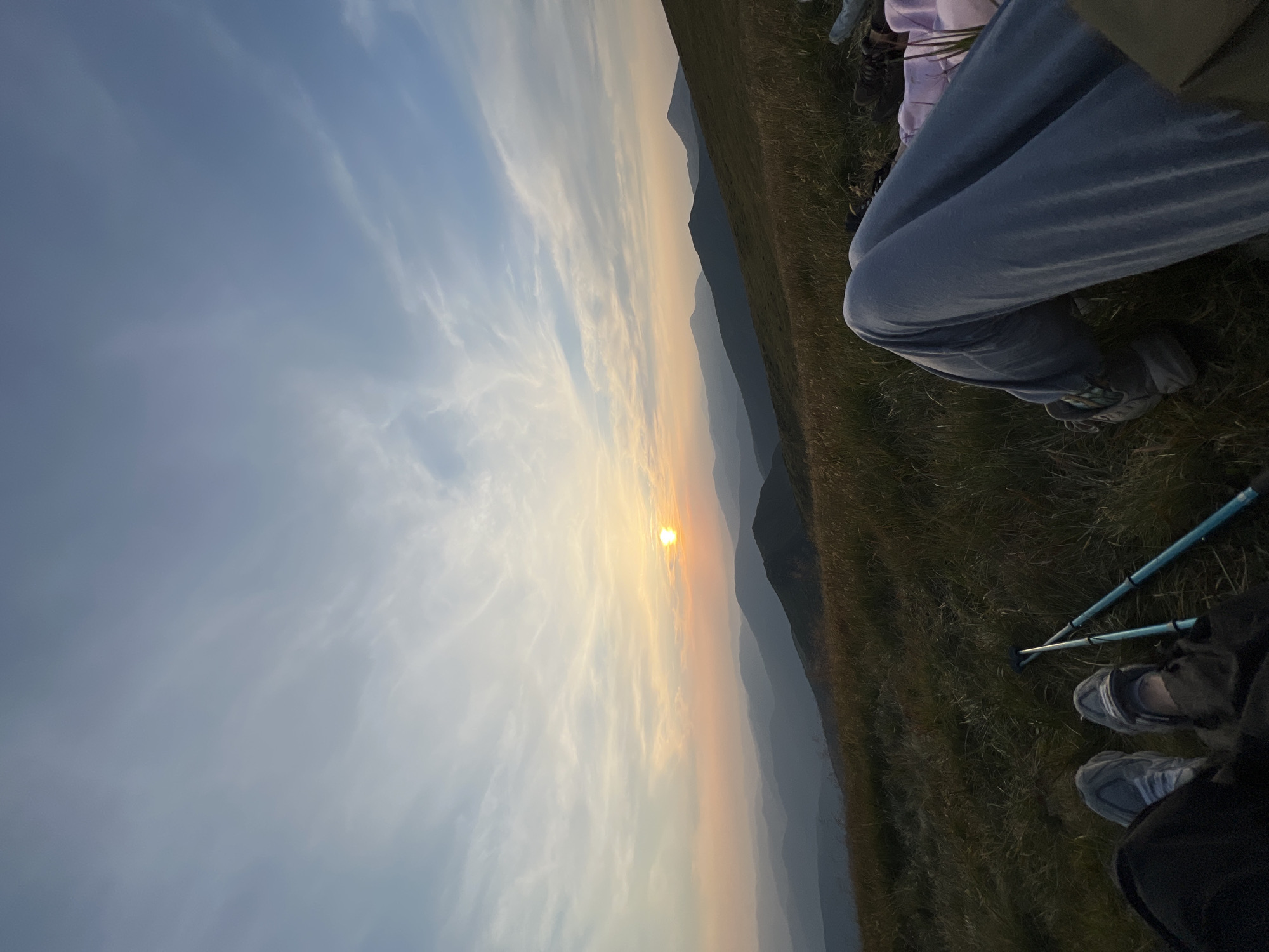 The image depicts a scenic mountain landscape at sunset, likely in Luhy, Ukraine. The foreground shows two pairs of legs and feet, suggesting two individuals are seated or resting on a grassy slope. One person wears grey athletic shoes and dark pants, while the other wears light grey athletic shoes and light blue sweatpants. Two blue hiking poles are visible between them.
The mid-ground features rolling hills covered in dry grass and some green vegetation. The background is dominated by layers of silhouetted mountain ranges fading into a hazy, orange and pink sky. The sun, a bright orb, is low on the horizon, casting a warm glow and illuminating wispy clouds. The overall atmosphere is serene and tranquil, indicative of a moment of rest during an outdoor excursion.