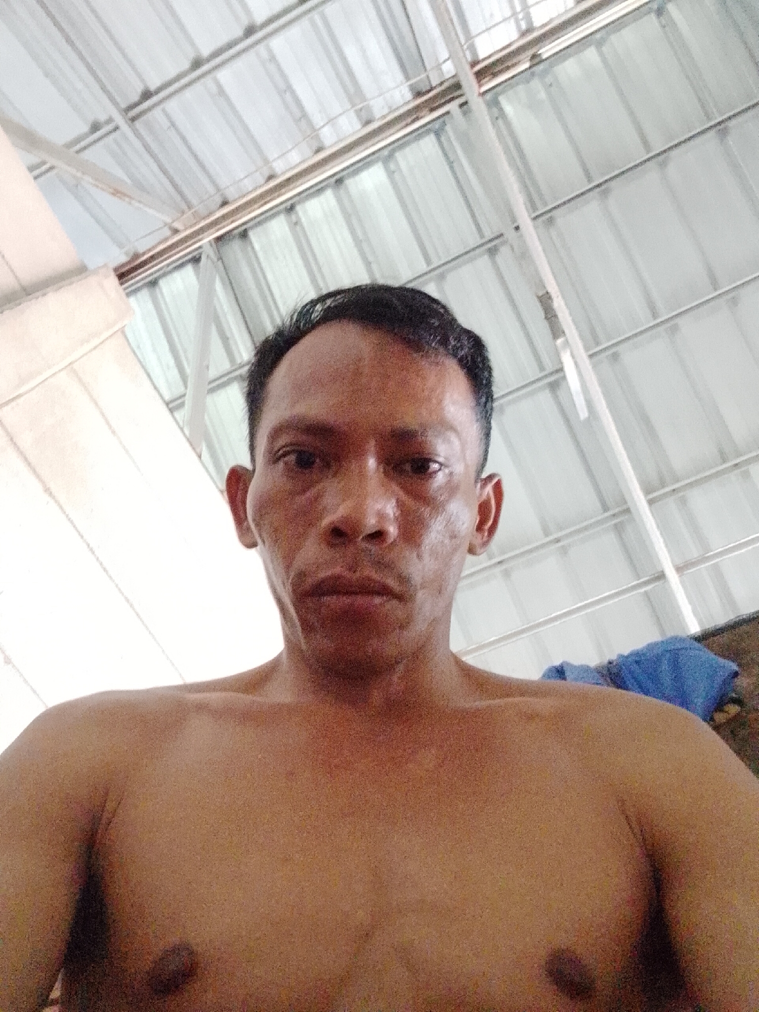 A man with short, dark hair is looking directly at the camera. He is shirtless. The background is an indoor setting with a metal roof structure visible. A blue object is visible behind his right shoulder. The city and state are unknown.