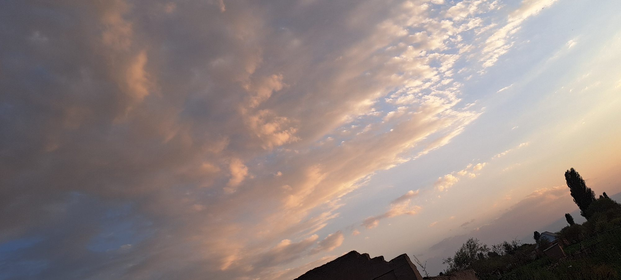 The image depicts a sky scene over Bash-Kaiyndy, Kyrgyzstan, during what appears to be sunset or sunrise. The sky is filled with clouds, varying in color from dark gray and blue on the left to lighter shades of peach, pink, and white towards the right, suggesting the sun's rays are illuminating them from the side. The clouds are arranged in streaks and patches, with some appearing wispy and others more dense.
In the lower portion of the image, the silhouette of a rooftop is visible on the far left, suggesting a building. To the right, the landscape transitions to a grassy hillside with trees. A tall, slender tree with dark foliage stands prominently in the middle right, with smaller trees and some structures, possibly houses, nestled amongst the vegetation further to the right. The overall environment appears rural or semi-rural, characterized by natural elements and modest human habitation. No people are visible in the frame. The perspective is looking upwards at an angle, with the horizon line obscured by the elevated viewpoint or the tilt of the camera.