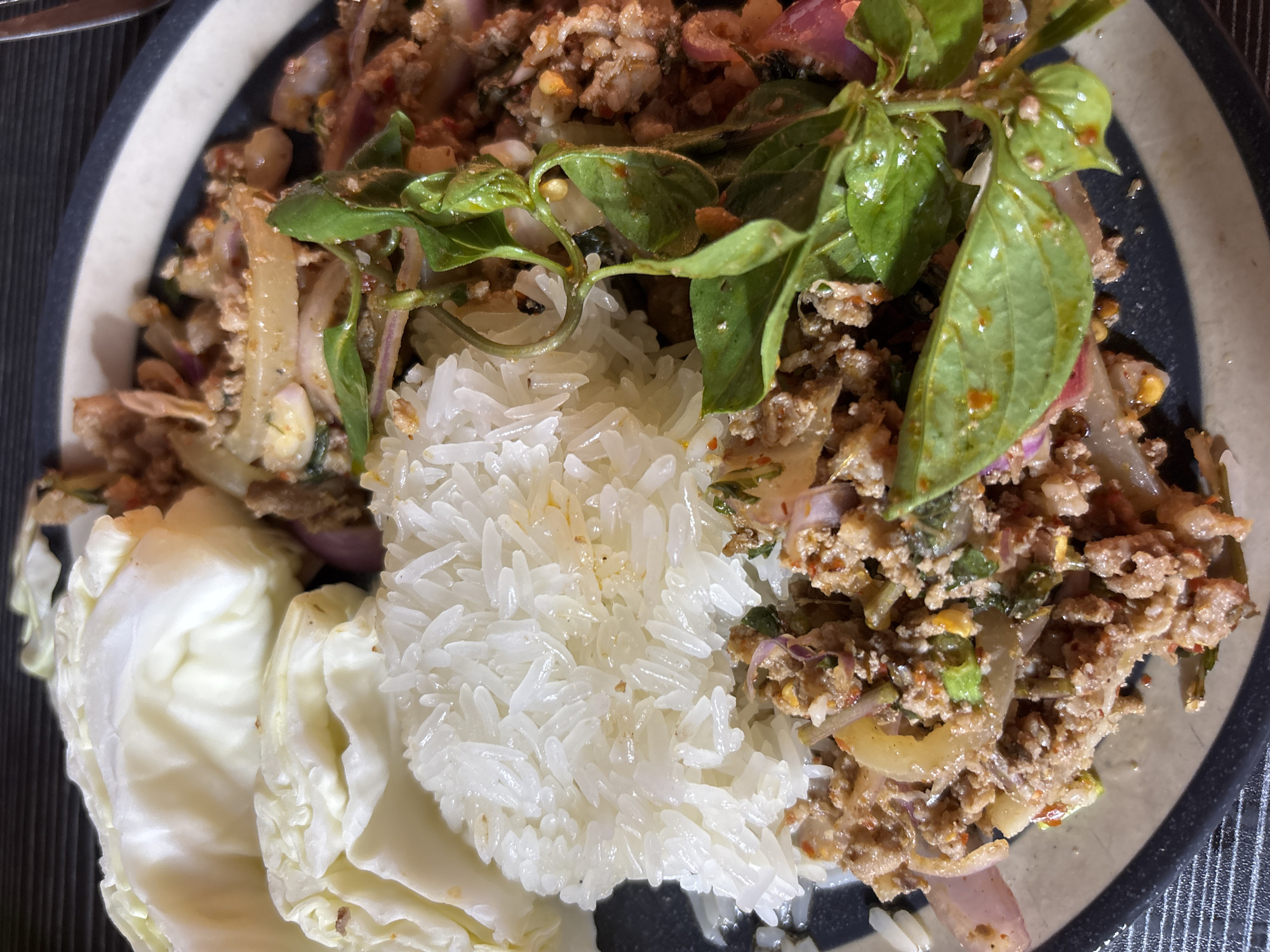 A plate of food is seen in a close-up shot. The dish appears to contain Larb, a type of minced meat salad popular in Southeast Asian cuisine, alongside a serving of sticky rice. Accompanying the Larb are some cabbage leaves. The meal is presented on a plate with a blue and white patterned rim. The location is likely บ้านบางรักษ์, Thailand, given the culinary context. No people are visible.
