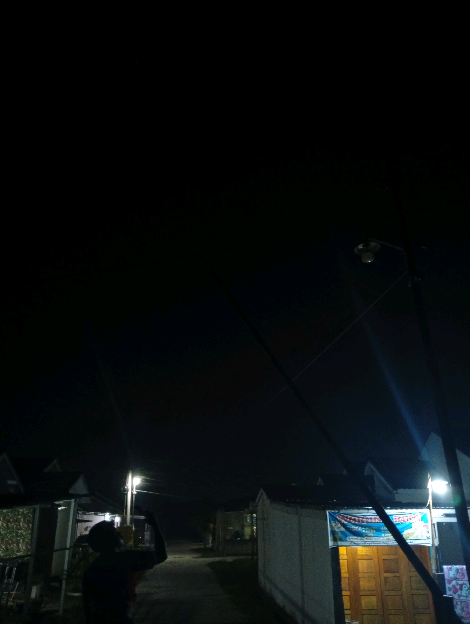 The image depicts a dimly lit street scene at night in Kualu, Indonesia. In the foreground, a silhouetted figure stands facing away from the camera, with their arm raised. To the right of the figure, a street lamp illuminates the scene, casting long shadows. In the background, several small buildings are visible, one of which has a banner displayed on its facade. The overall impression is of a quiet, residential area at night.
