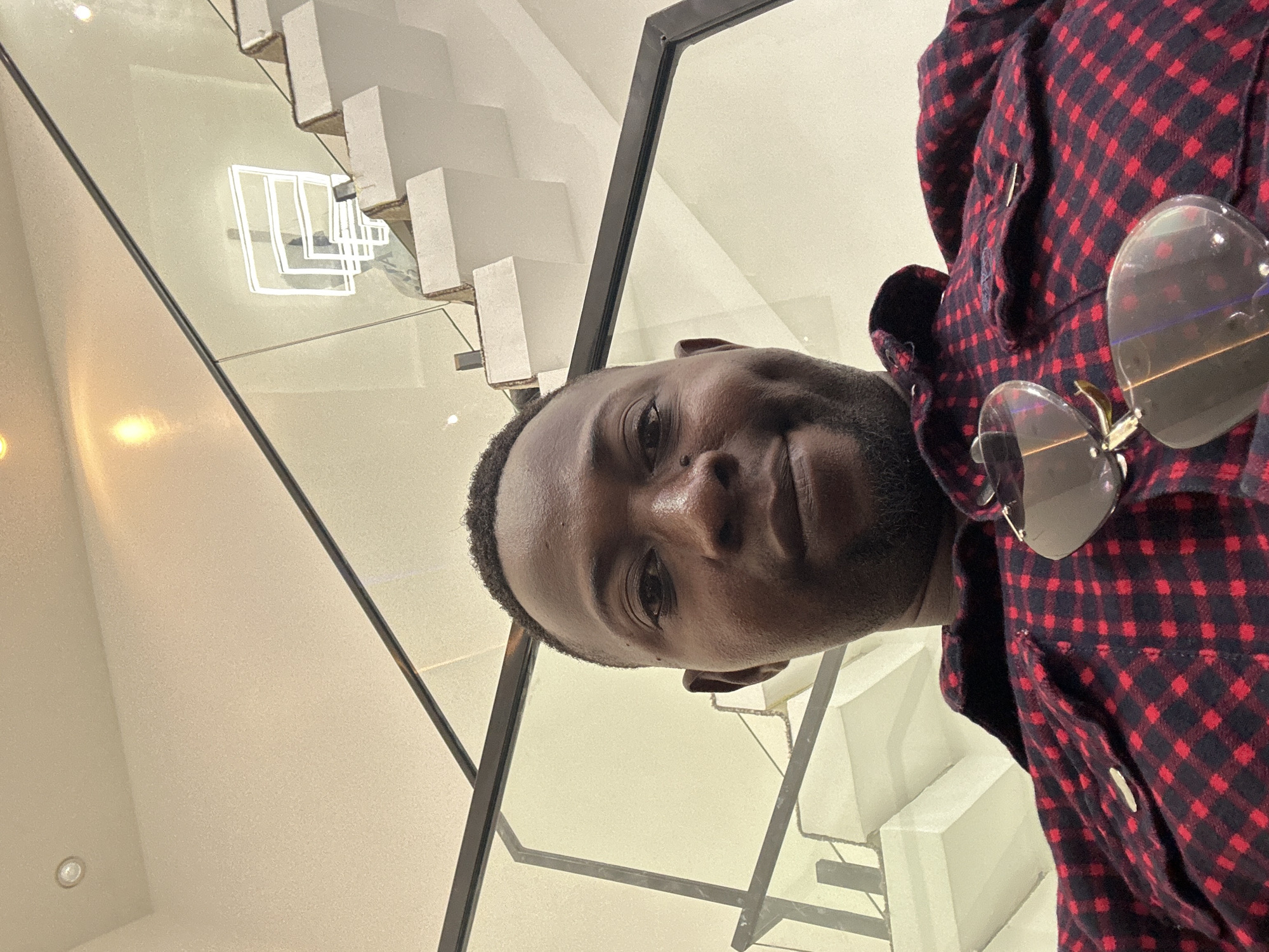 A close-up selfie shows a man wearing a red and black plaid shirt. He is wearing large, round sunglasses pushed up on his head. The background is a modern interior with a glass railing and a staircase. The lighting suggests it is daytime. The overall mood is casual and relaxed. The image appears to be taken in Unguwar Sarki, Nigeria.