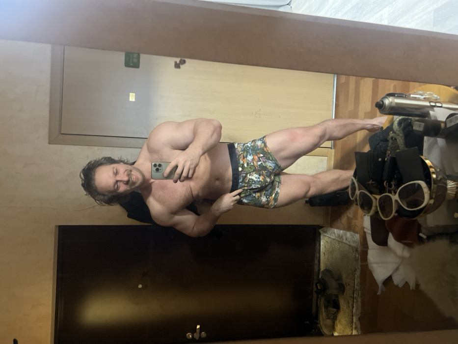 A shirtless man poses in front of a mirror, holding a phone to take a selfie. He wears printed shorts and has a mustache. The background includes a door, a beige wall, and a messy shelf with sunglasses, a helmet, clothes, and other items. The image appears to be rotated, presenting a tilted perspective.