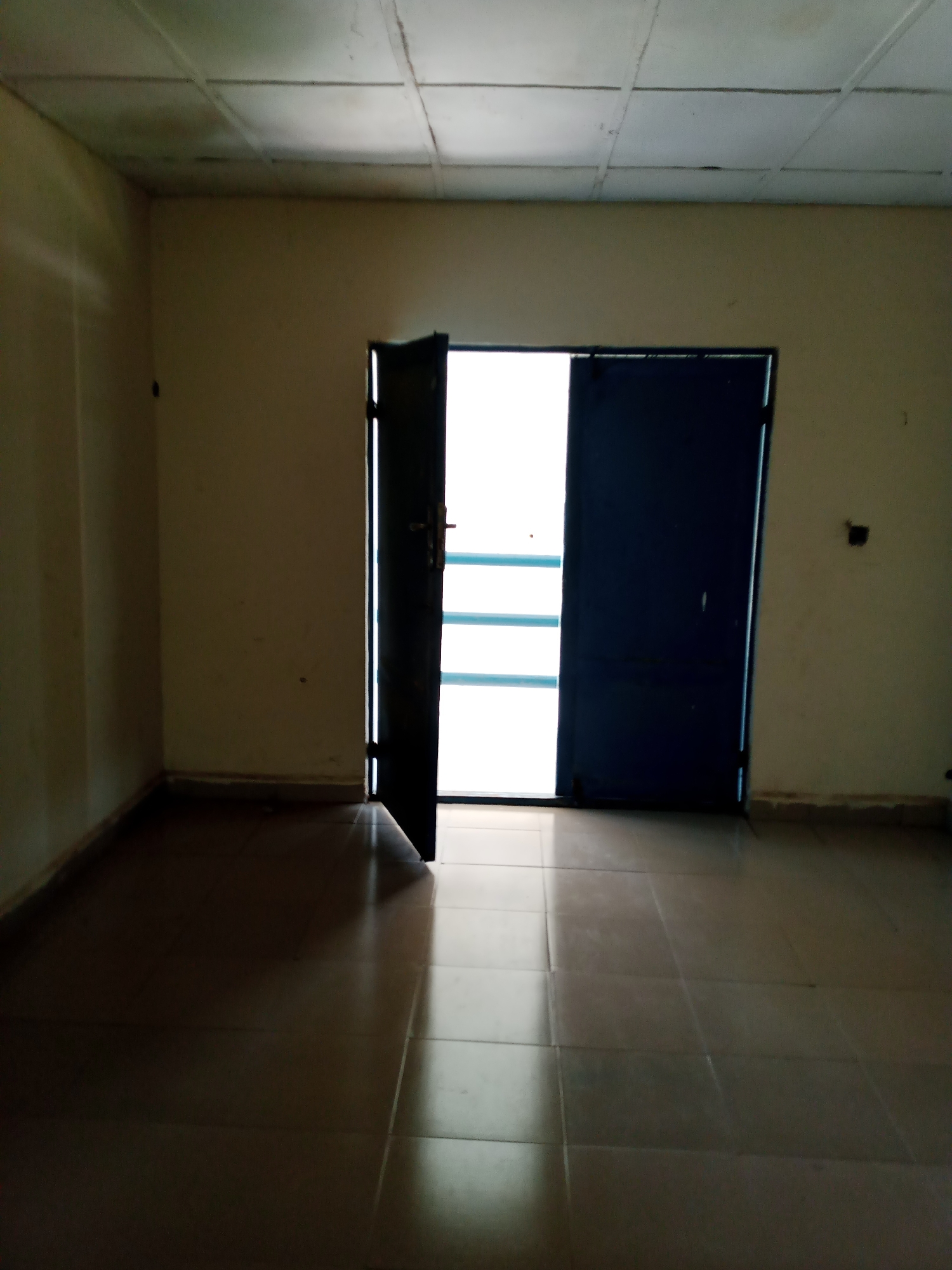 The image shows the interior of an empty room with tiled floors and off-white walls. There is a double door, one leaf of which is open, revealing a brightly lit area outside. The door is dark blue and has a metal frame. The ceiling appears to be made of acoustic tiles. The room is dimly lit, with shadows cast by the open door. The overall impression is of a simple, utilitarian space. The scene is located in Jalingo, Nigeria, within a residential or commercial building.