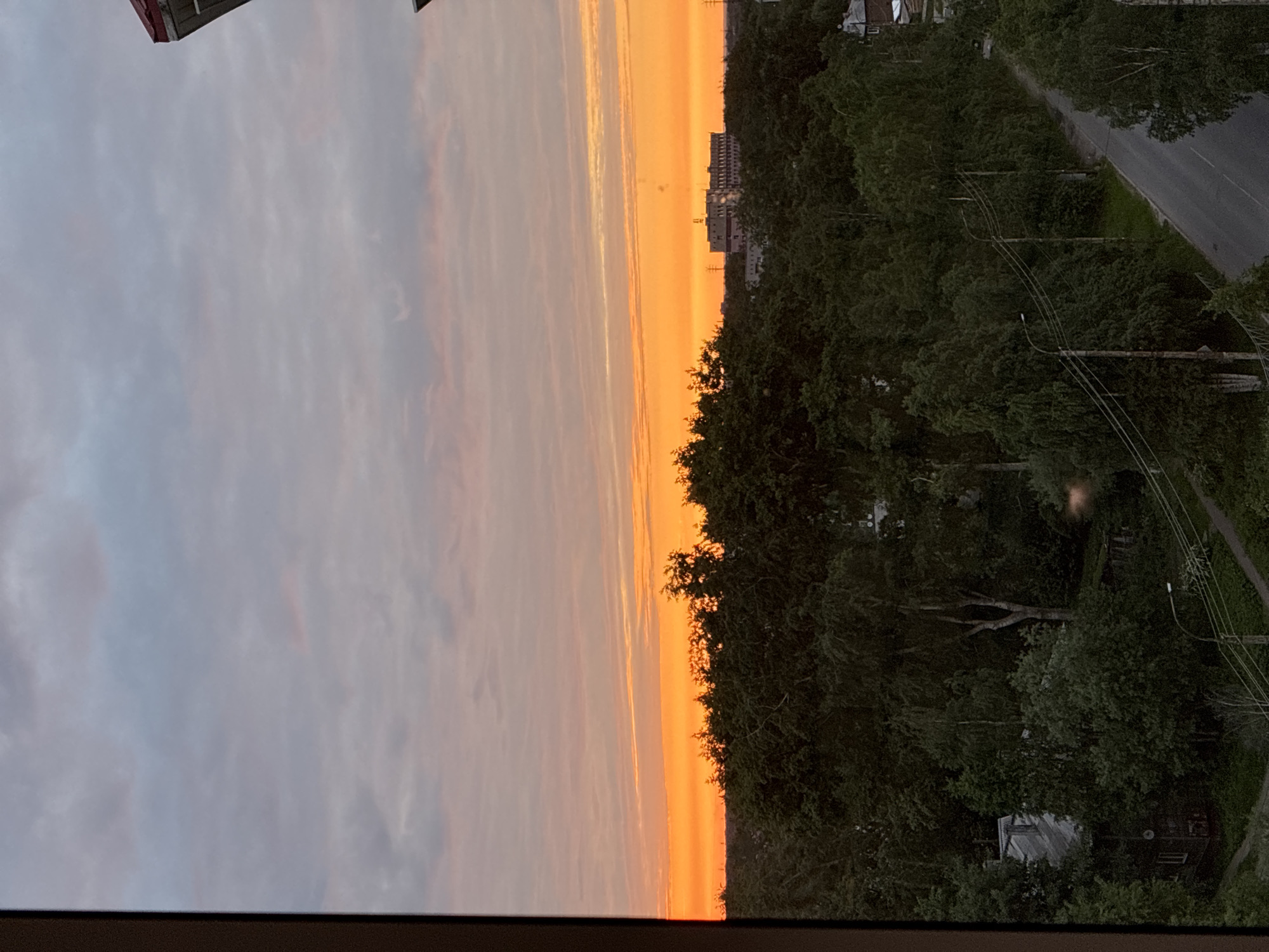 An elevated, outward-facing view captures a sunset or sunrise over a landscape in Unknown, Unknown. The sky transitions from a light grey with scattered clouds at the top to an intense band of vibrant orange and yellow at the horizon, marked by horizontal cloud formations. Below this bright horizon, a dense, dark green forest canopy dominates the midground. In the far distance, a large, multi-story building with a flat roof and visible structures stands partially silhouetted against the bright sky, with faint utility poles also visible. In the foreground, a paved road winds diagonally from the lower right, bordered by utility poles carrying multiple wires and equipped with streetlights. To the lower left, partially obscured by trees, a multi-story wooden house with a grey gabled roof is visible. Additional shrubbery and dirt paths are present around the road and structures. A dark frame edge from a window or opening is visible on the far left and partially on the top right. No individuals are present in the scene.