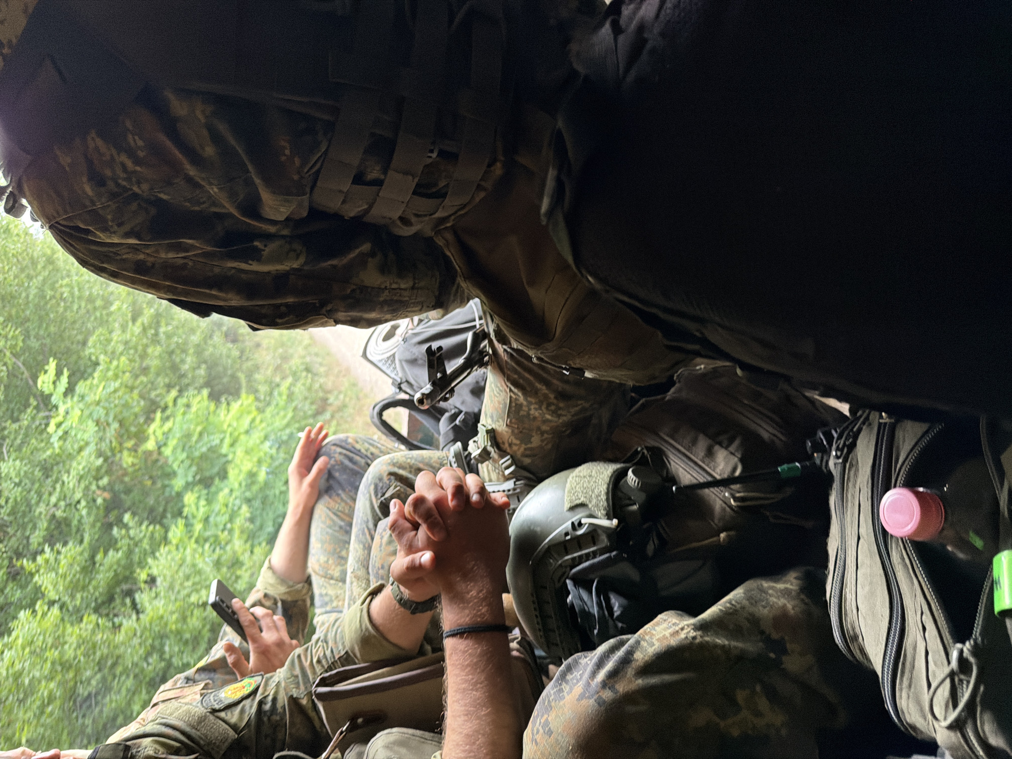 The image shows a group of individuals in military-style camouflage uniforms seated in what appears to be the back of a vehicle. The environment is outdoors, with lush green trees and foliage visible in the background, suggesting a rural or wooded setting. The individuals are equipped with gear including helmets, tactical vests, and bags. One person is holding a smartphone, while another has their hands clasped. The scene is captured from a low angle, looking upwards and towards the back of the vehicle. There is no visible indication of the location being Beliakovets, Bulgaria.