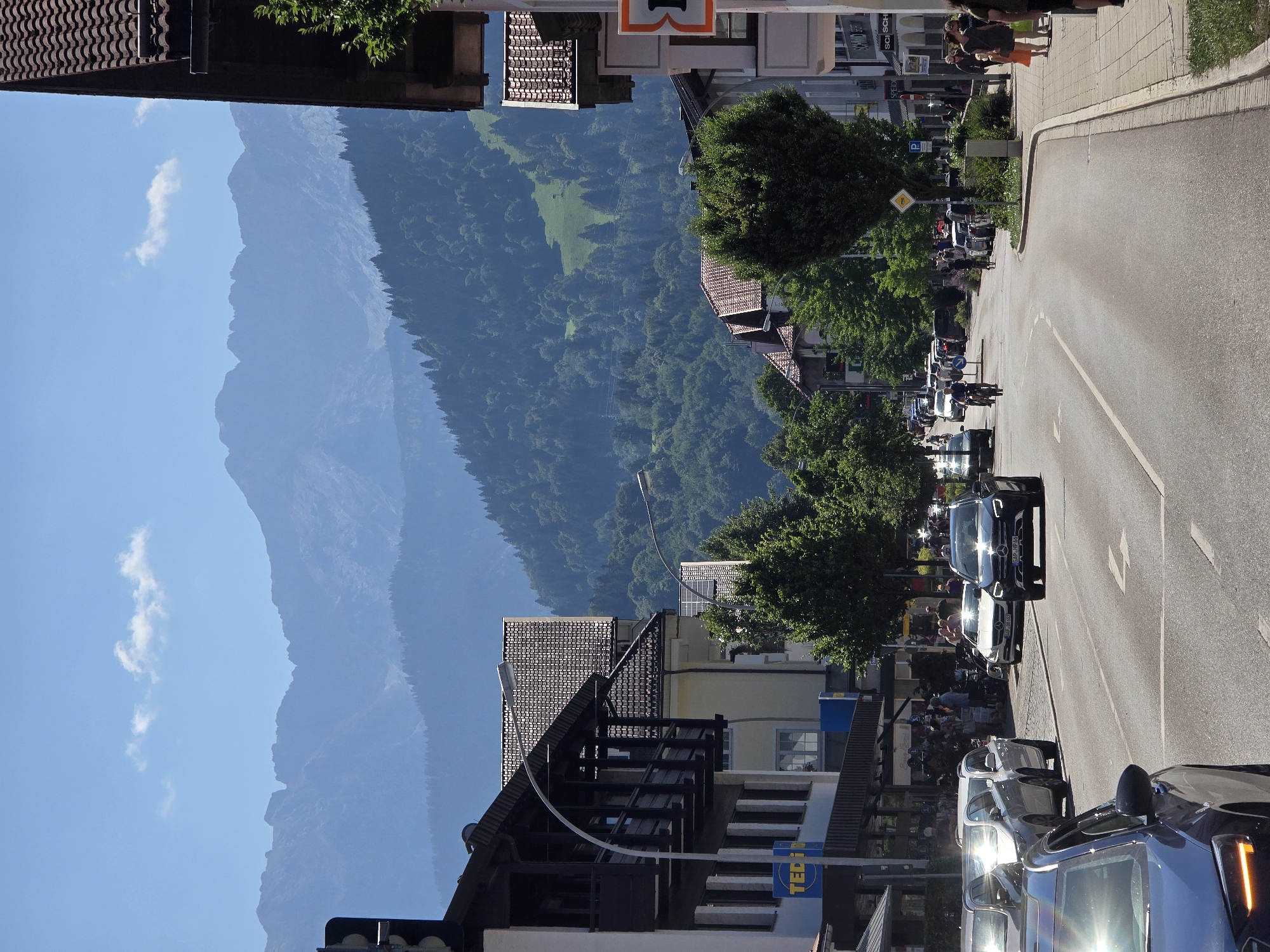 The image depicts a street scene in Garmisch-Partenkirchen, Germany, with a backdrop of majestic mountains. The foreground features a two-lane road with several cars driving and parked. On the left side of the road, a building with a dark wooden balcony overlooks the street. A blue sign with "TEDI" is visible on this building. On the right side, there are more buildings with shops and pedestrians walking on the sidewalk.
In the middle ground, lush green trees line the street, and more buildings with tiled roofs are visible. The road curves gently to the right, leading the eye towards the distant mountains. The mountains themselves are a prominent feature, with rugged peaks and slopes covered in dense green forest, ascending towards a clear blue sky with a few wispy clouds. The overall atmosphere is that of a sunny day in a picturesque European town nestled in a mountainous region.