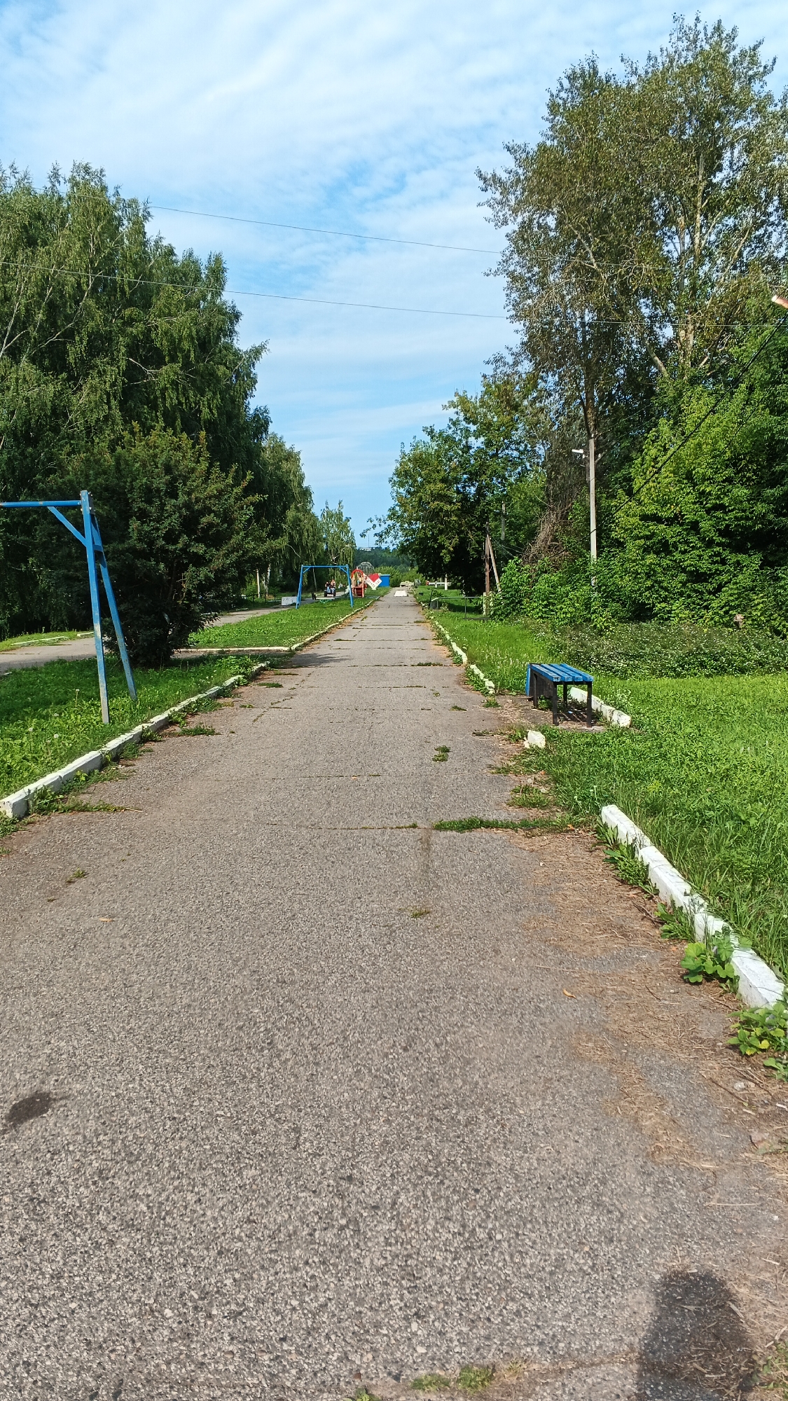 A paved pathway stretches into the distance, flanked by grassy areas and trees. On the left, a blue metal swing set is visible. Further down the path, on the right, a blue metal bench rests on a concrete base. The sky is partly cloudy with a few wispy white clouds against a blue background. The scene appears to be a park or a recreational area in the city of Podgornoye, Russia. The pathway shows signs of wear, with some cracks and patches of dry grass growing through. The foliage is lush and green, suggesting a summer or late spring setting. There are no people visible in the image. Overhead power lines are present in the upper portion of the frame.