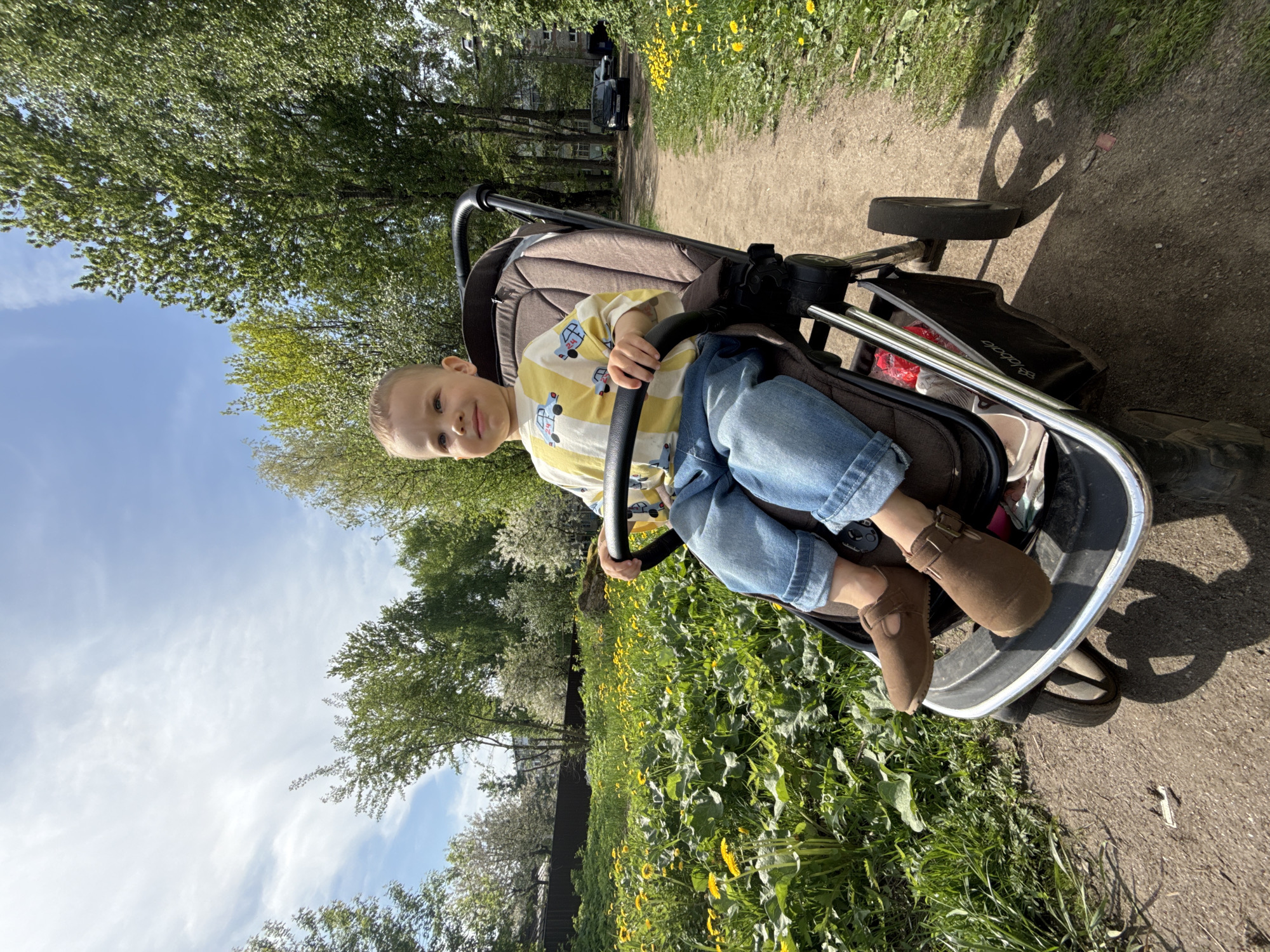 The image shows a young child sitting in a stroller on a path surrounded by greenery, potentially in Rusanovo, Russia. The child is wearing a yellow and white striped shirt with car designs, blue jeans, and brown shoes. The stroller is positioned on a dirt path bordered by grass and yellow wildflowers. Trees are visible in the background, with a building and a dark-colored vehicle further down the path. The sky is partially visible with white clouds.