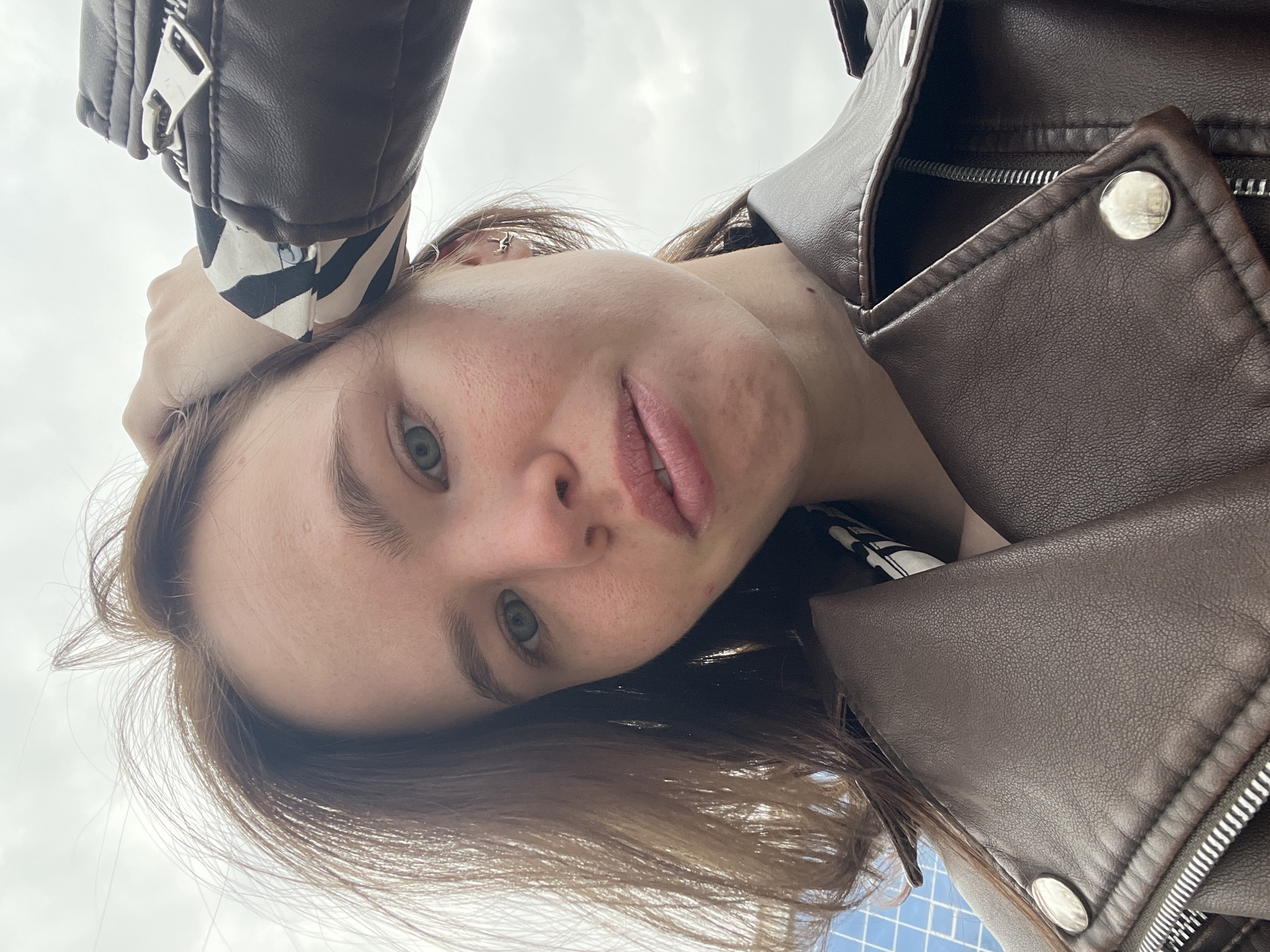The image is an upward-angled, rotated selfie of a young woman, taken in Chaikovsky, Russia. Her face is in the center, looking directly at the camera with a neutral to slightly pensive expression. She has light brown hair, light-colored eyes (blue/green), and full lips.
She is wearing a brown leather-like biker jacket, distinguished by silver zippers and prominent snap buttons. A black and white striped garment is visible beneath the jacket's sleeve and collar. A small, silver-colored earring is noticeable in her left ear. Her left hand is raised, partially obscured by her jacket sleeve, likely holding the camera that captured the shot.
The background consists mainly of a bright, overcast sky, suggesting a daytime setting with diffuse natural light. In the very bottom right corner, a subtle blue and white patterned surface is visible, possibly indicating a tiled area or ground. No visible text is present in the image.