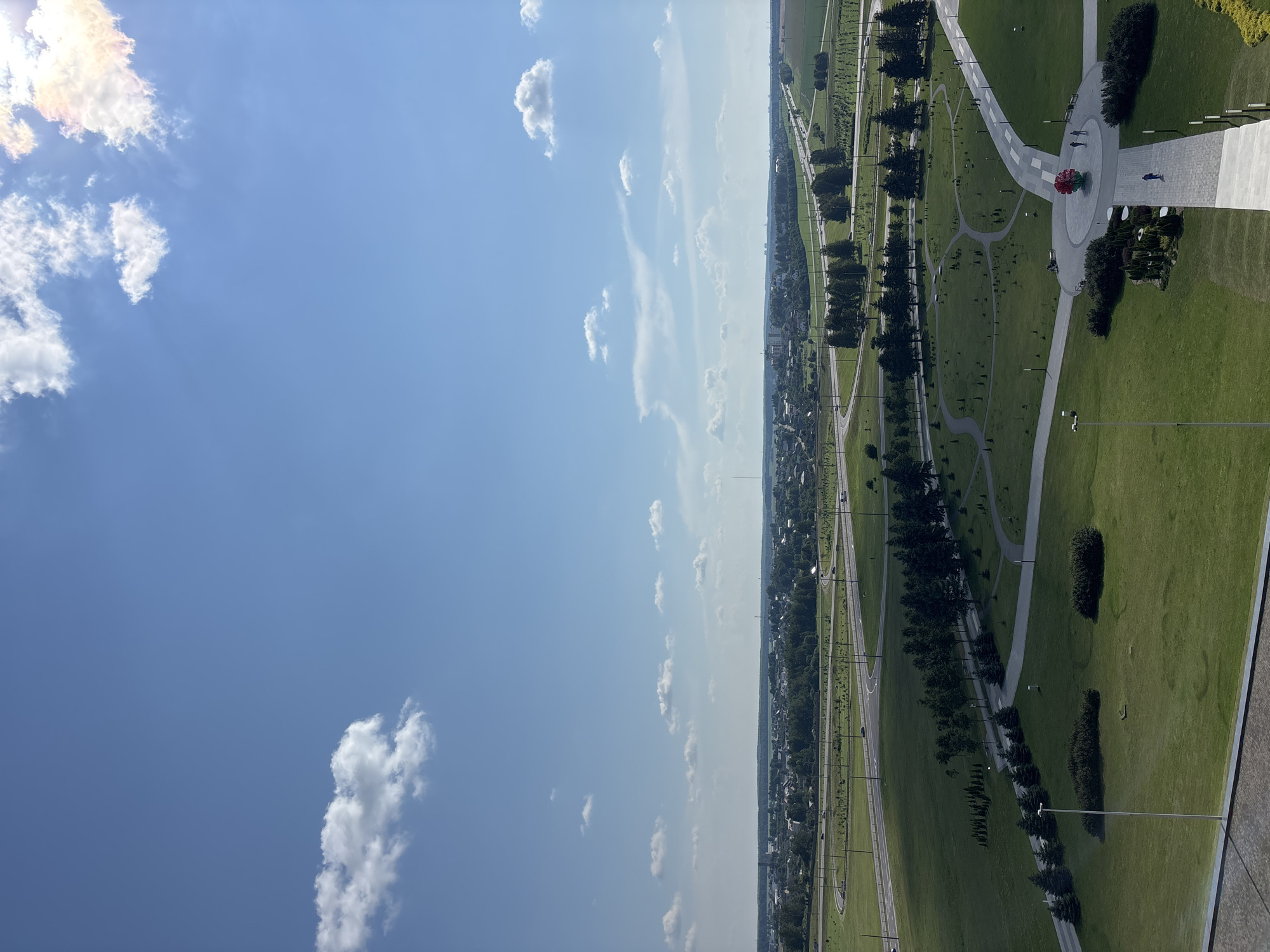 An aerial view captures a modern park area within the city of Ozyaritska-Slabadski rural council, Belarus. A bright blue sky with scattered white clouds dominates the upper portion of the image, with the sun glinting from the top right. Below, a vast expanse of green lawn is intersected by paved pathways, creating a park-like setting. Several people are visible walking along these paths. A prominent, large red sculpture, resembling a stylized flower, is situated at a central intersection of pathways. Adjacent to the park, a complex highway interchange with multiple lanes and curves is visible, with cars traversing the roads. Further in the distance, a town or city with low-rise buildings and trees stretches across the horizon. The overall scene depicts a blend of urban infrastructure and recreational green space under a clear sky.